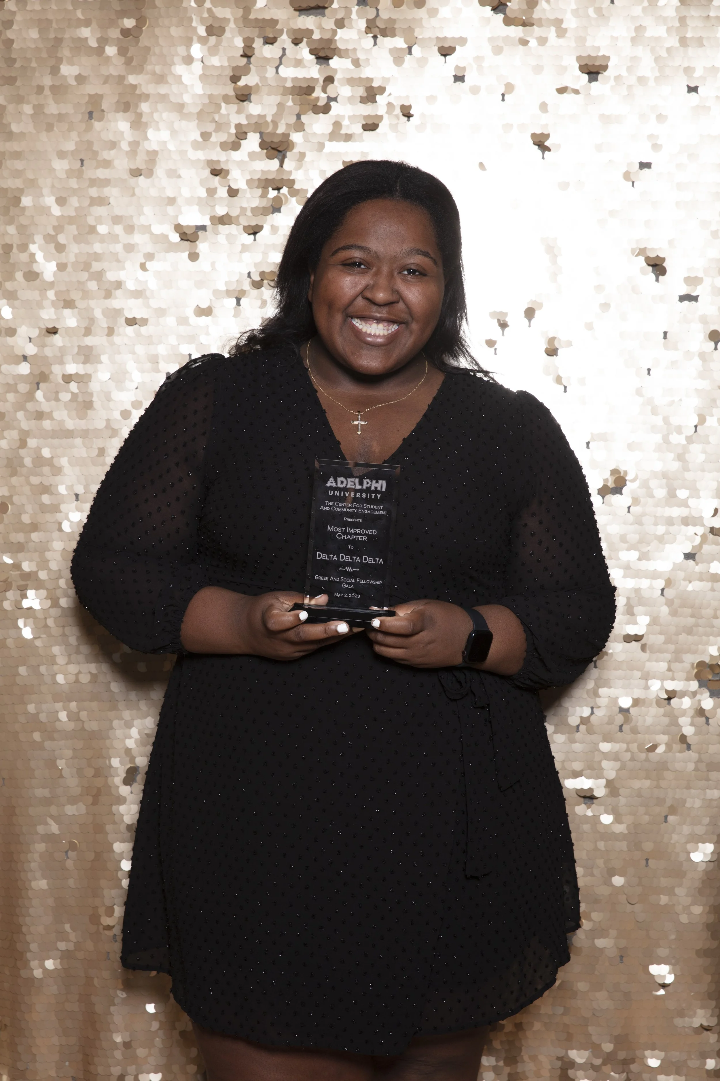 A young woman smiling and holding a trophy in front of a gold sequin backdrop. She is wearing a black dress with bubble sleeves, a cross necklace, and a smartwatch.