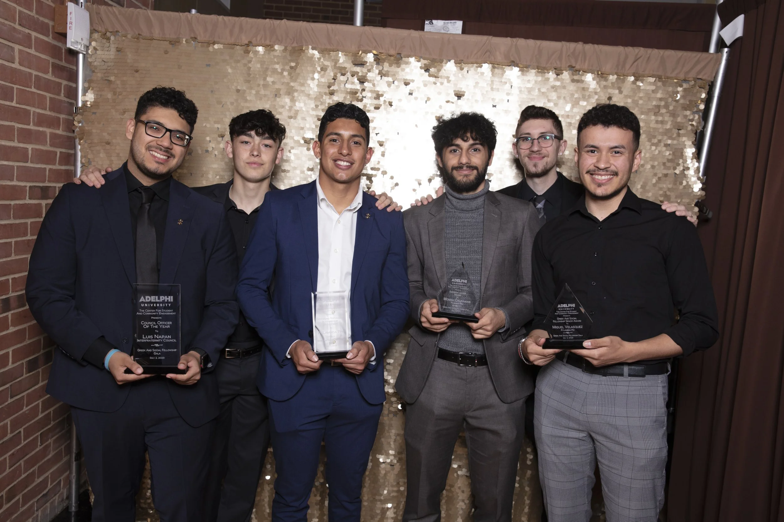 Six young men dressed in formal attire standing together at an awards ceremony, holding awards, smiling, in front of a gold sequin backdrop.