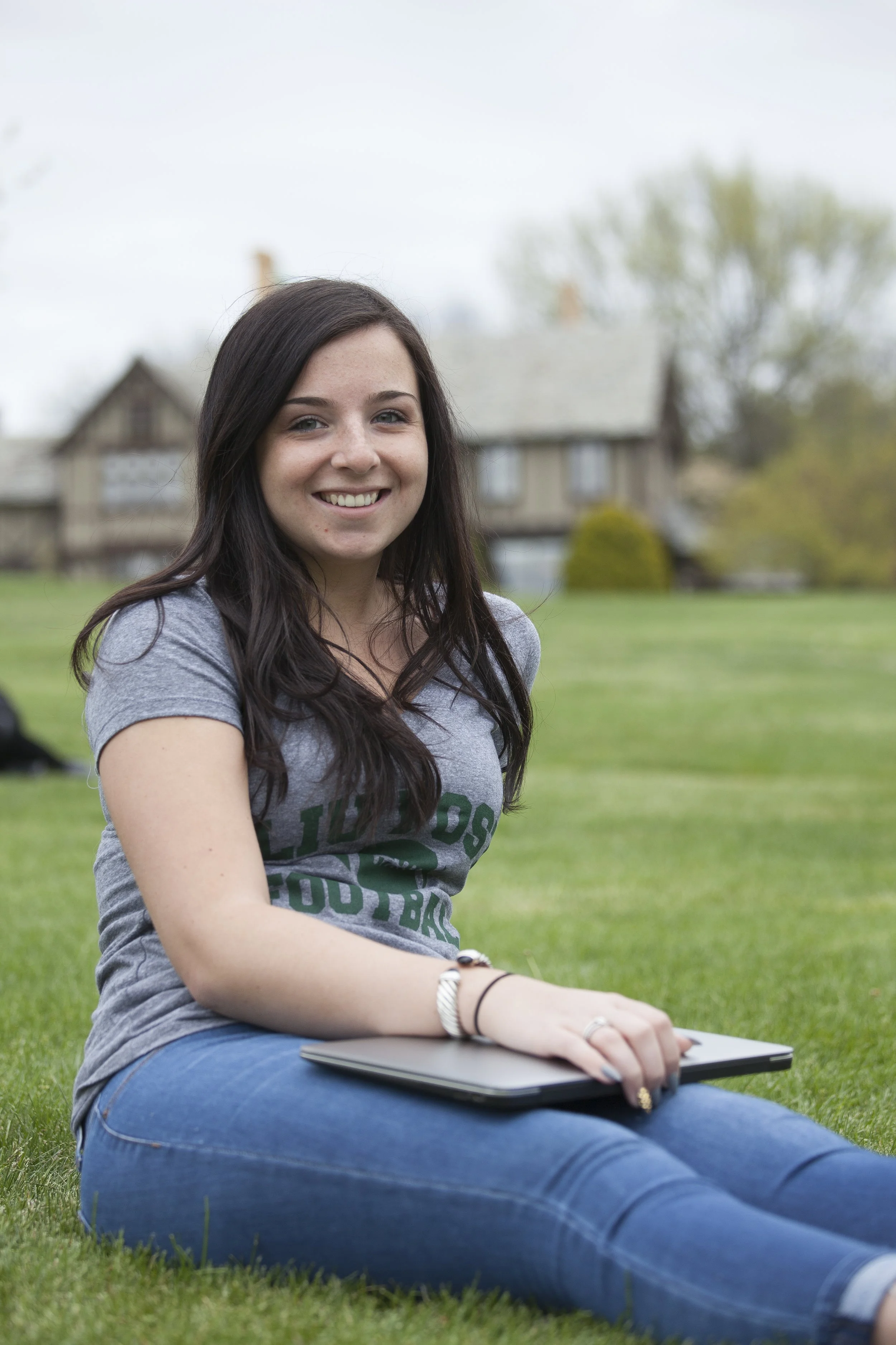 A young woman sitting on grass outdoors, smiling, holding a closed laptop on her lap, with a house and trees in the background.
