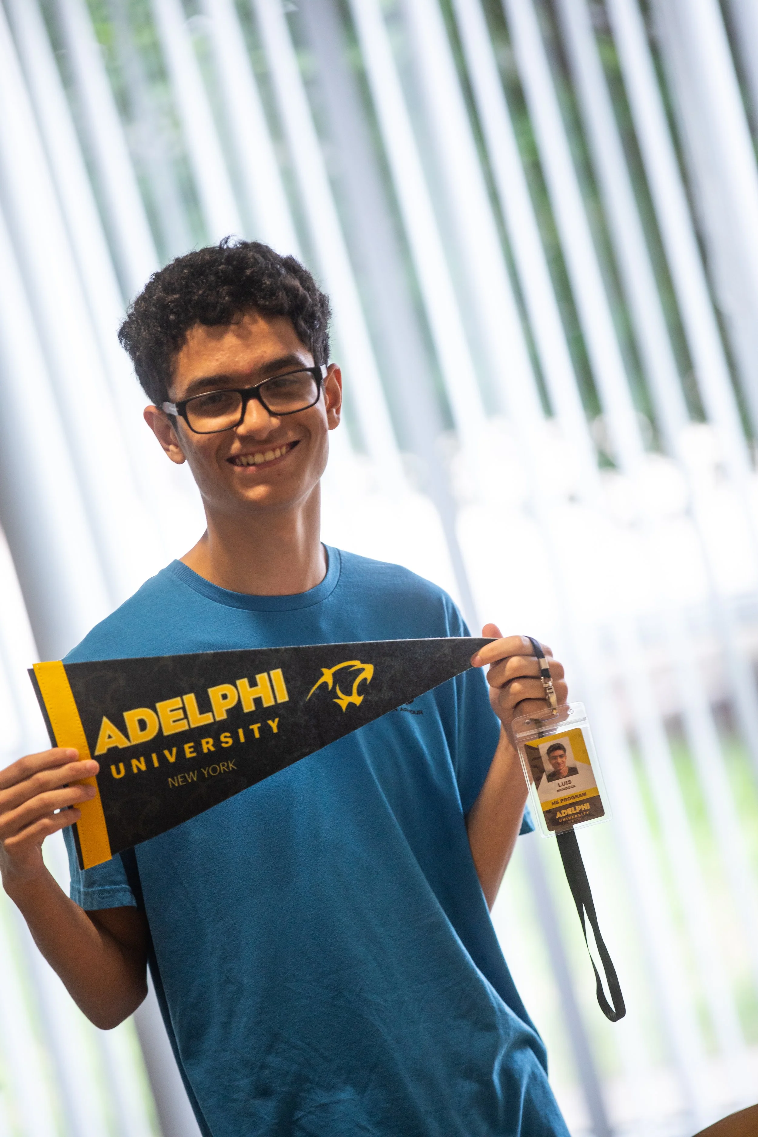 A young man with curly hair and glasses smiling while holding a black and yellow pennant for Adelphi University, standing in front of tall, vertical window blinds and holding an ID badge.
