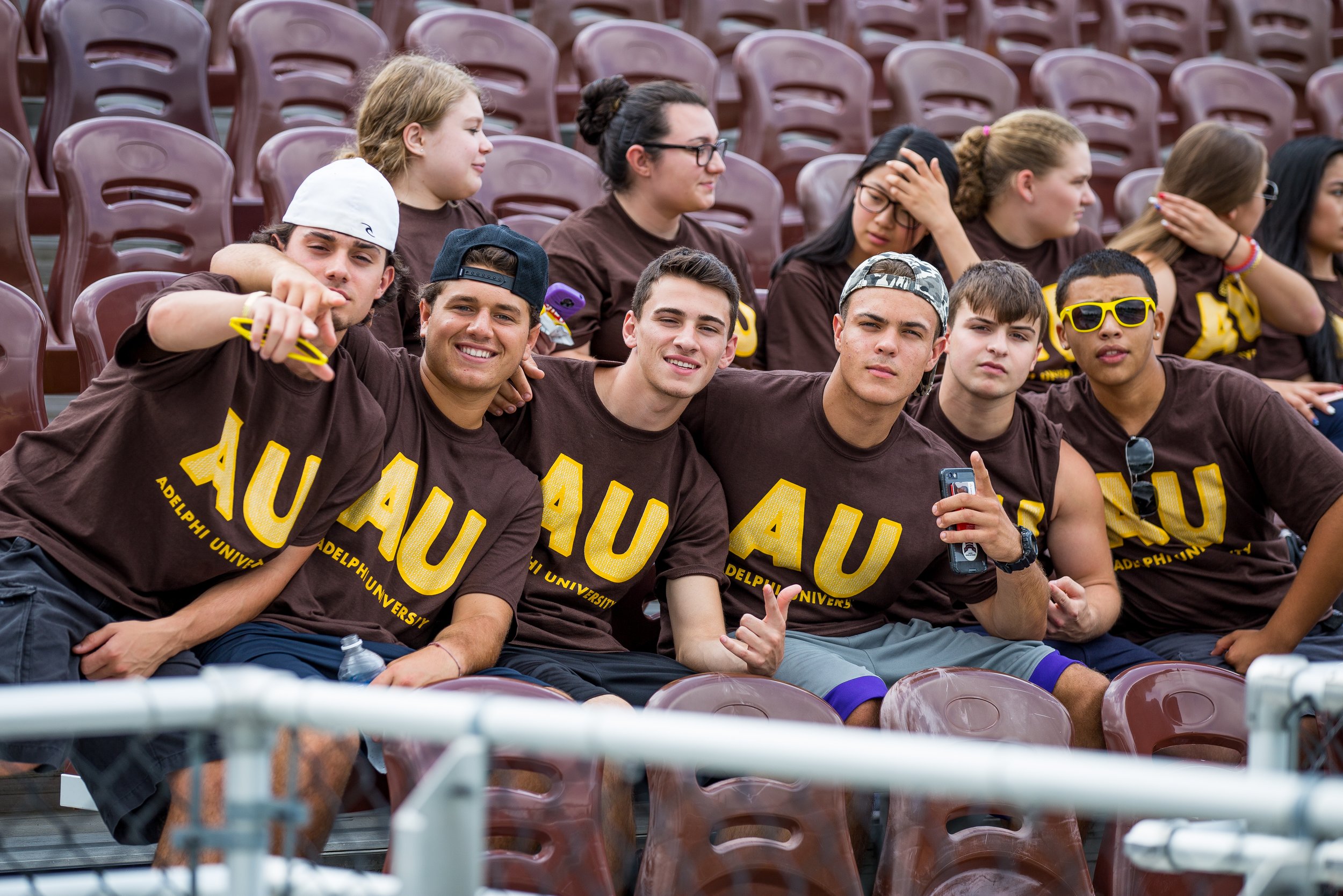 Group of college students sitting on stadium bleachers, wearing matching brown 'AU' shirts from Adelphi University, smiling and posing for the camera.