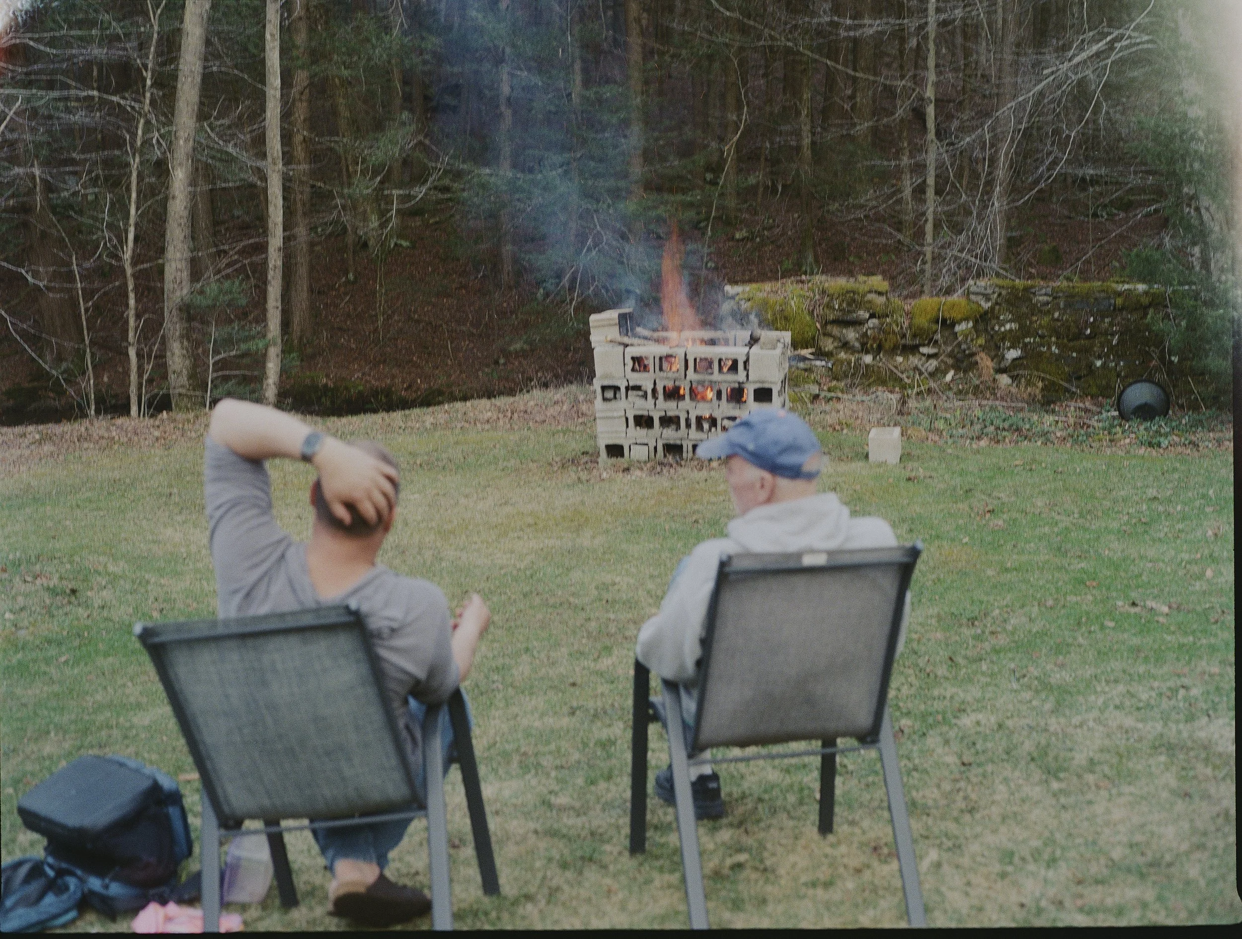 Two men sitting in outdoor chairs watching a small fire burning inside a cinder block structure in a wooded area.