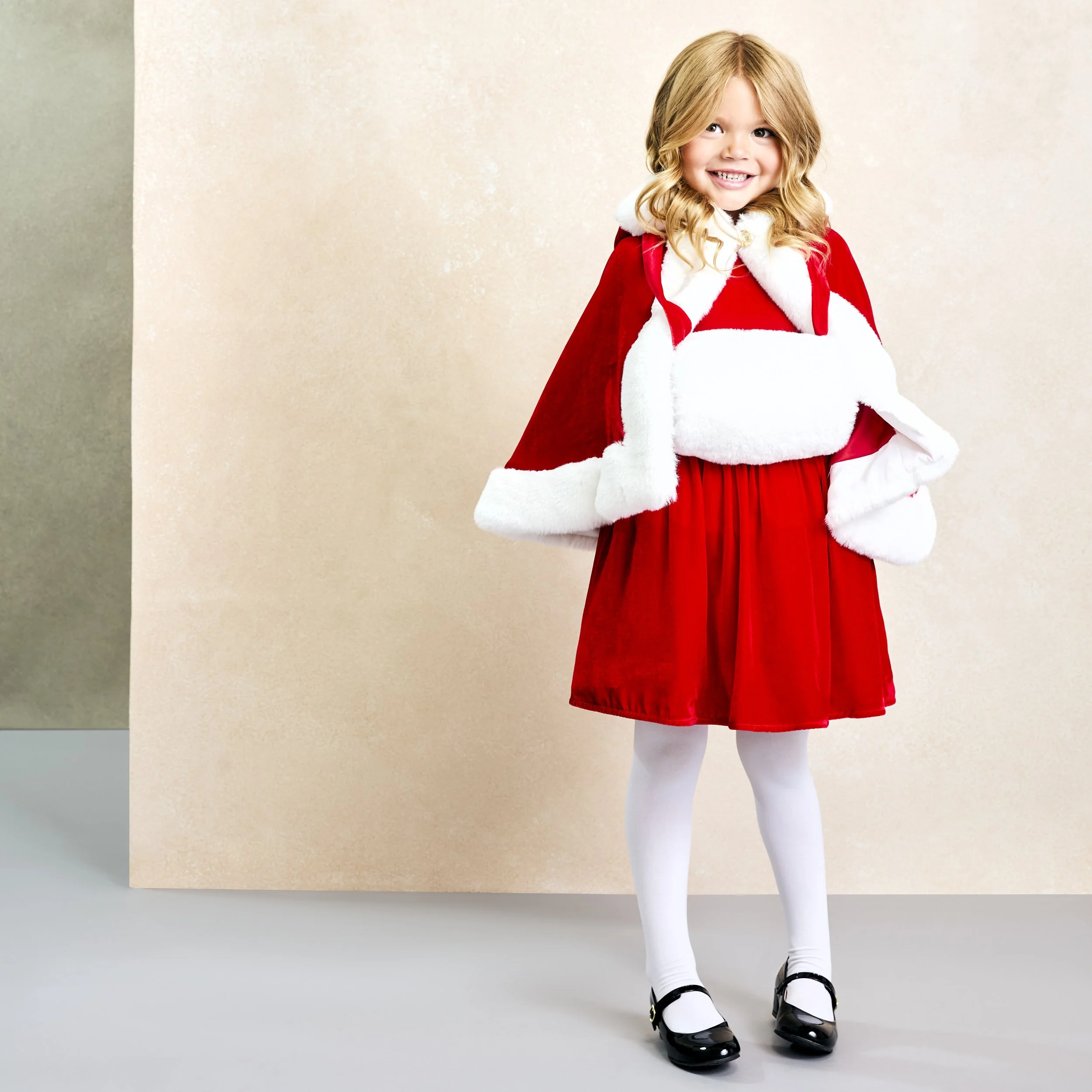 Young girl dressed in a festive red and white Christmas outfit with a cape, white tights, and black shoes, standing and smiling.