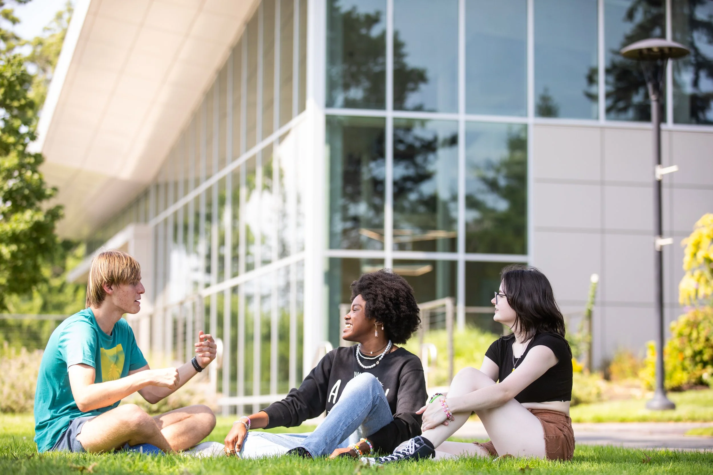 Three young people sitting on grass outside a modern building, engaged in conversation and smiling.