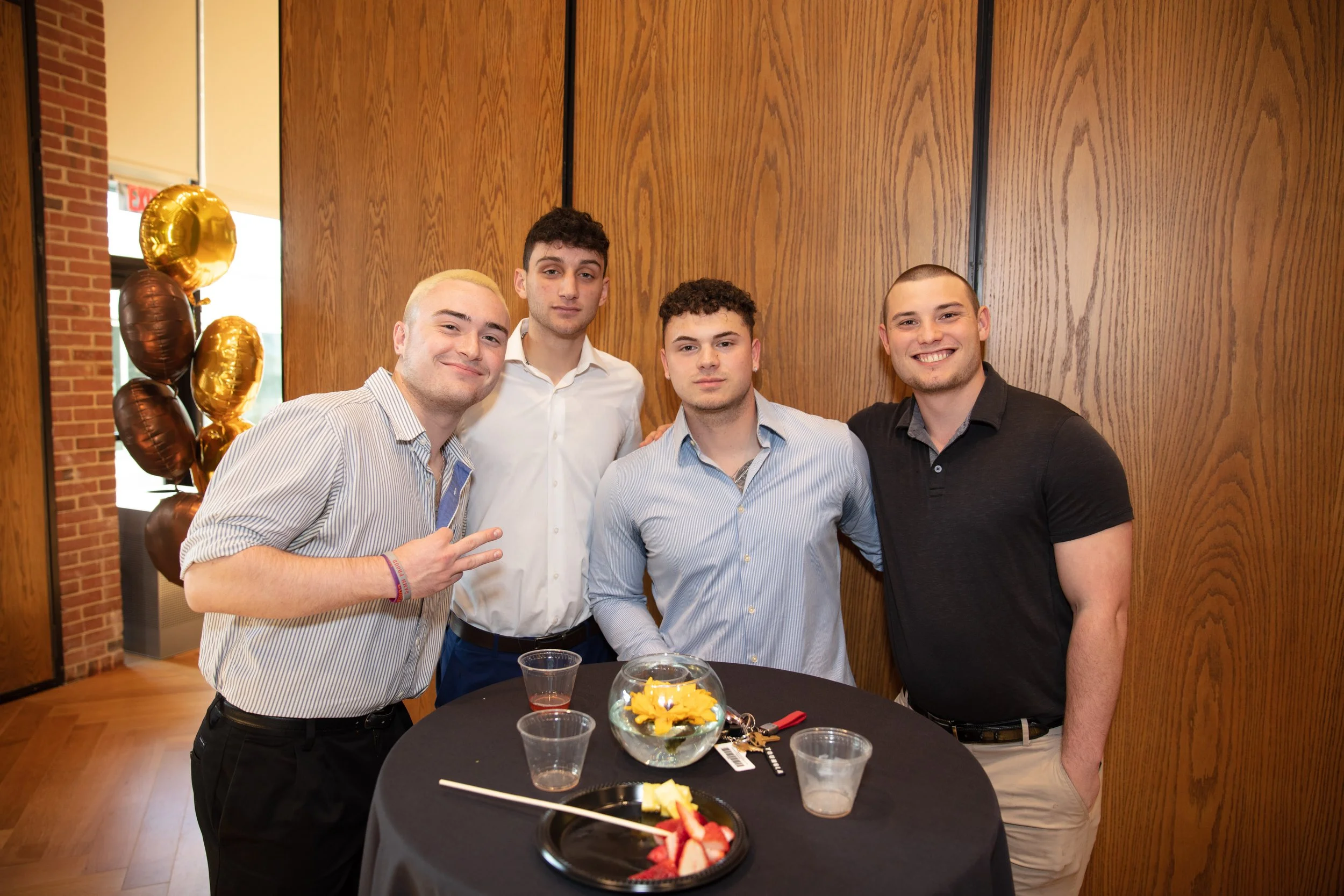 Four young men at a party, standing behind a table with food and drinks, smiling and making peace signs, with balloons in the background.