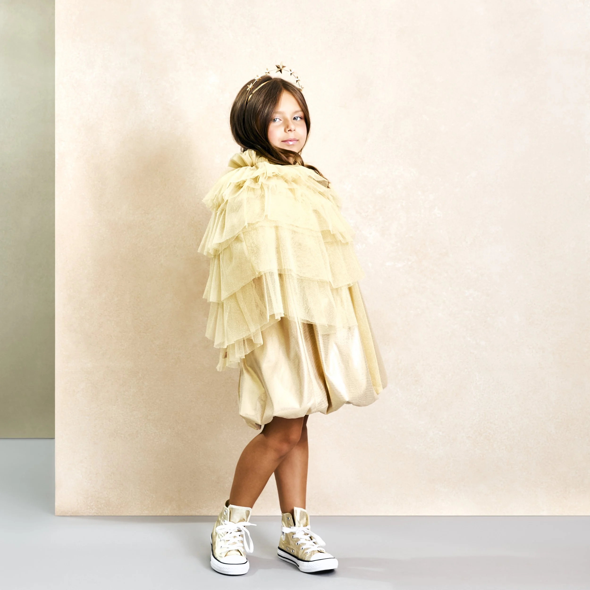 Girl wearing a gold tulle layered dress, gold high-top sneakers, and a star headband, standing against a beige background.