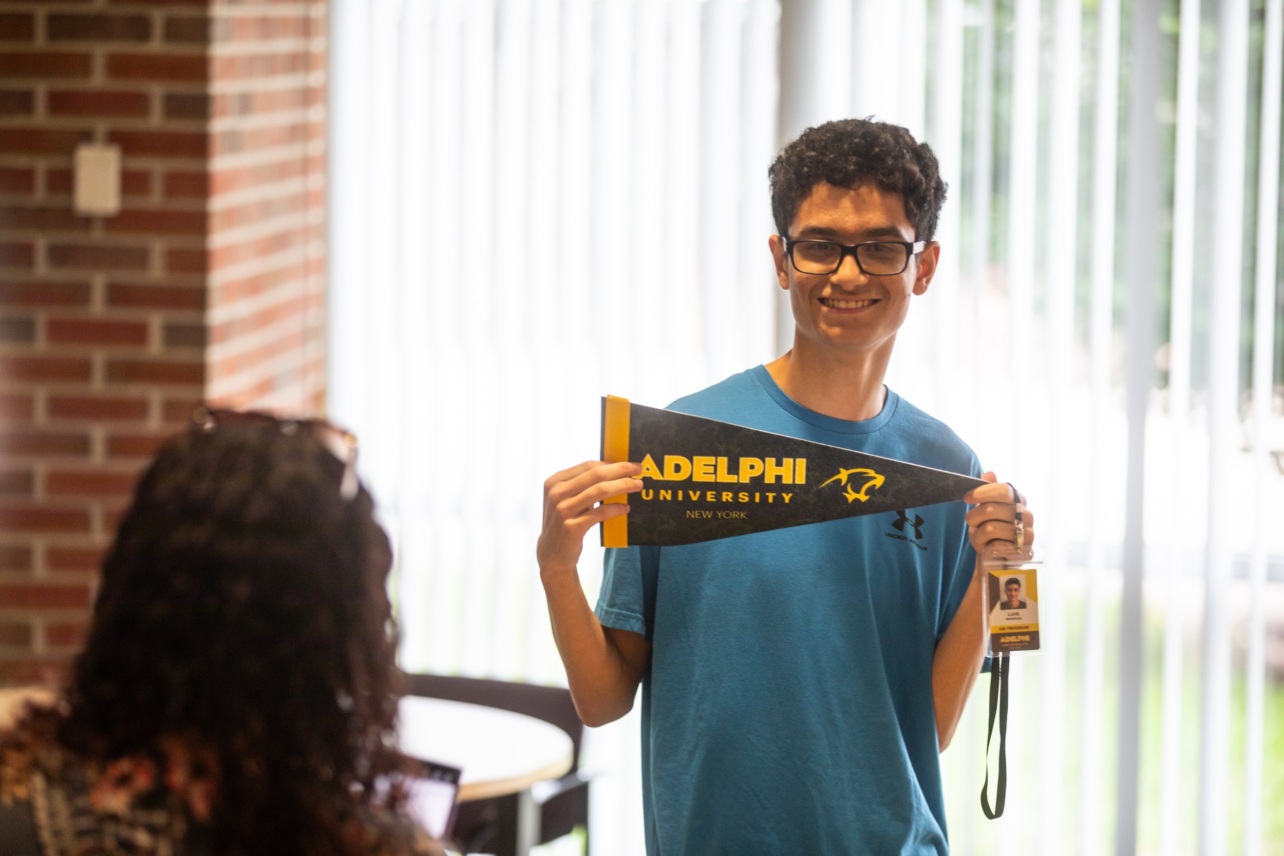 A young man with glasses smiling and holding a Pennant that reads 'Adelphi University New York' while standing inside a building with large windows and vertical blinds.