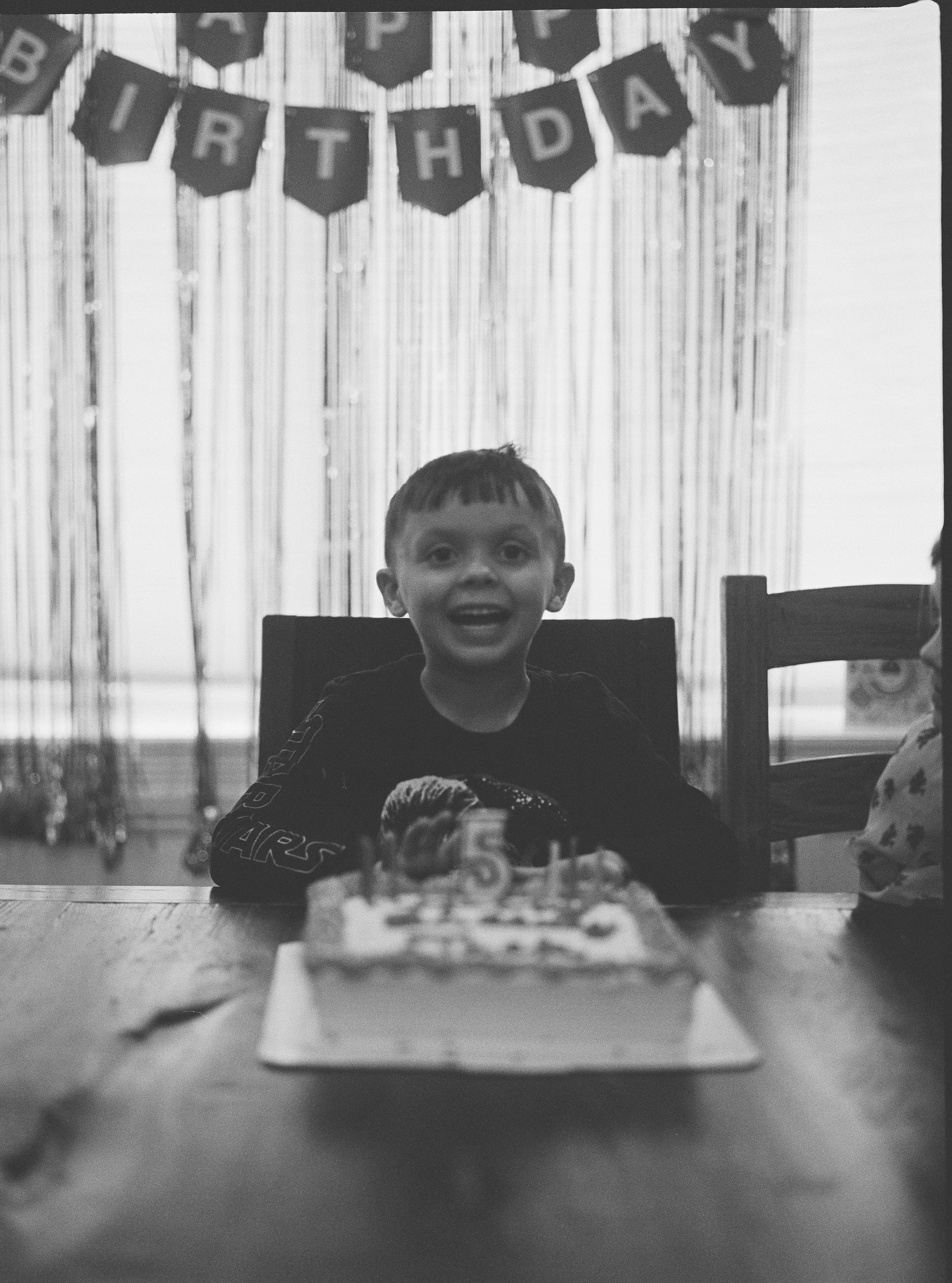 A young boy with short hair and a big smile sitting at a table in front of a birthday cake, with birthday banners hanging in the background.