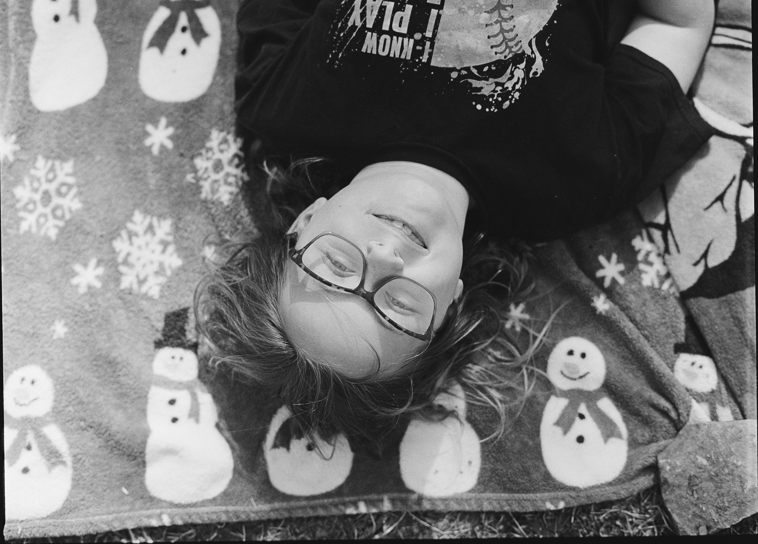 A young girl with curly hair and glasses lying on a carpet decorated with snowmen and snowflakes, smiling at the camera.