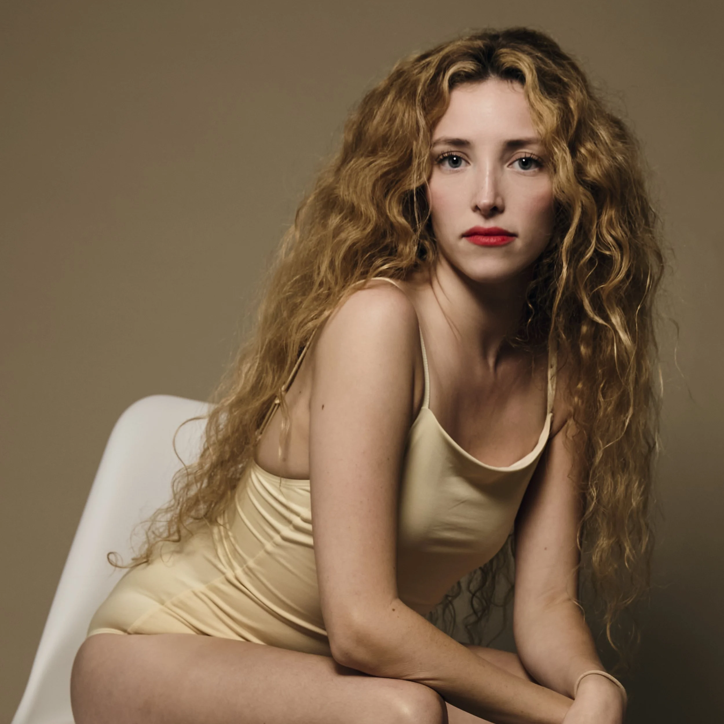 A young woman with long, curly red hair and light skin, wearing a beige satin slip dress, sitting on a white chair against a plain taupe-colored wall.