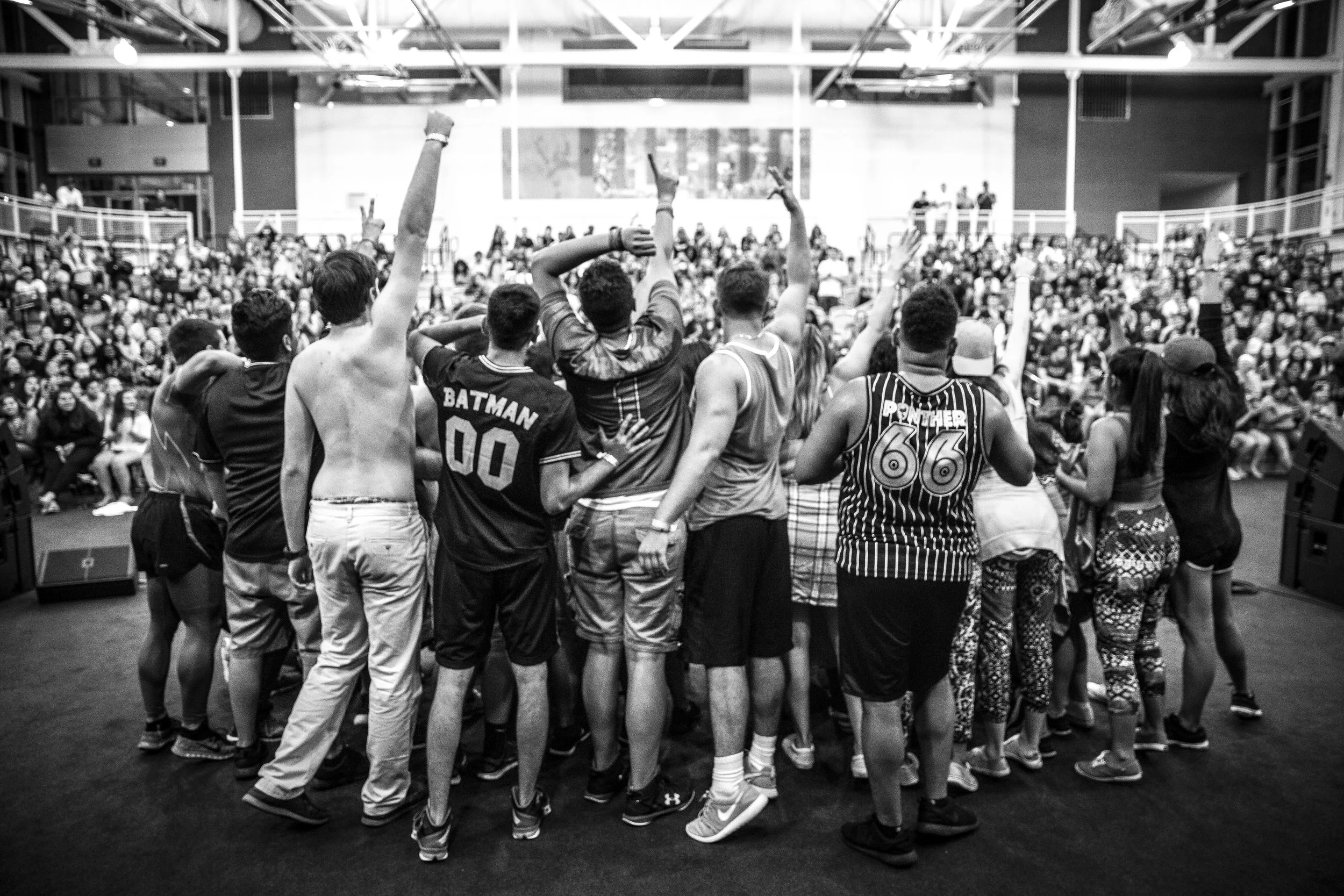 A large group of diverse people standing together in a gymnasium or event space, with a crowd sitting in the background, some raising their hands, and some wearing sports jerseys.