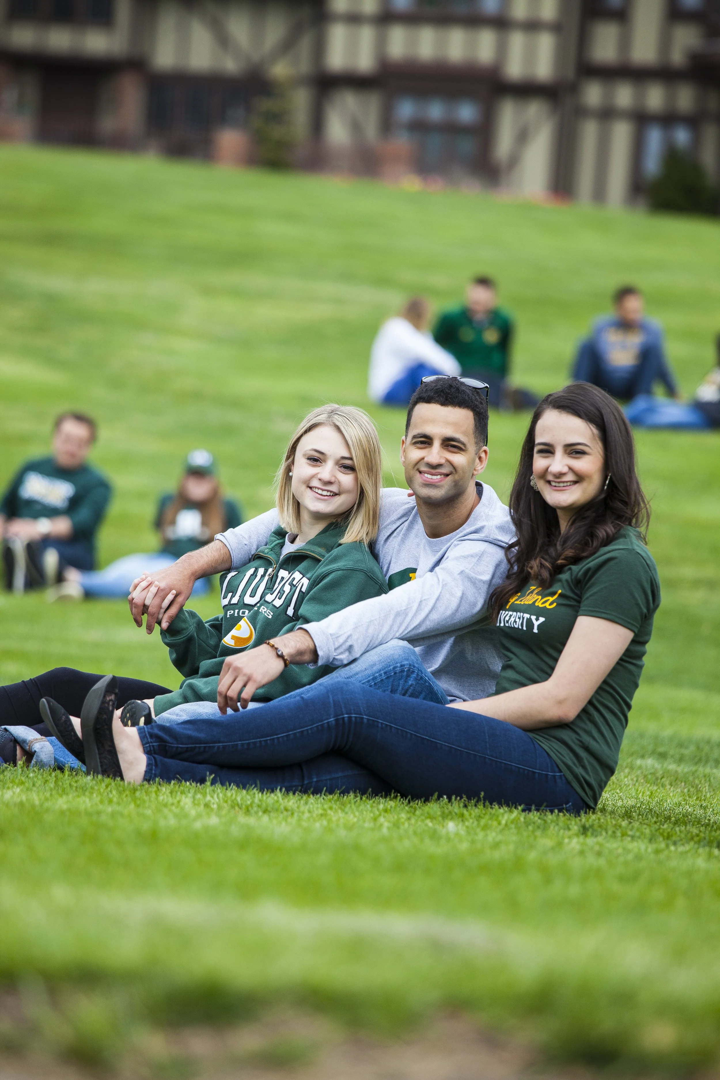 Three college students sitting on a grassy field, smiling at the camera, with other people sitting and relaxing in the background.