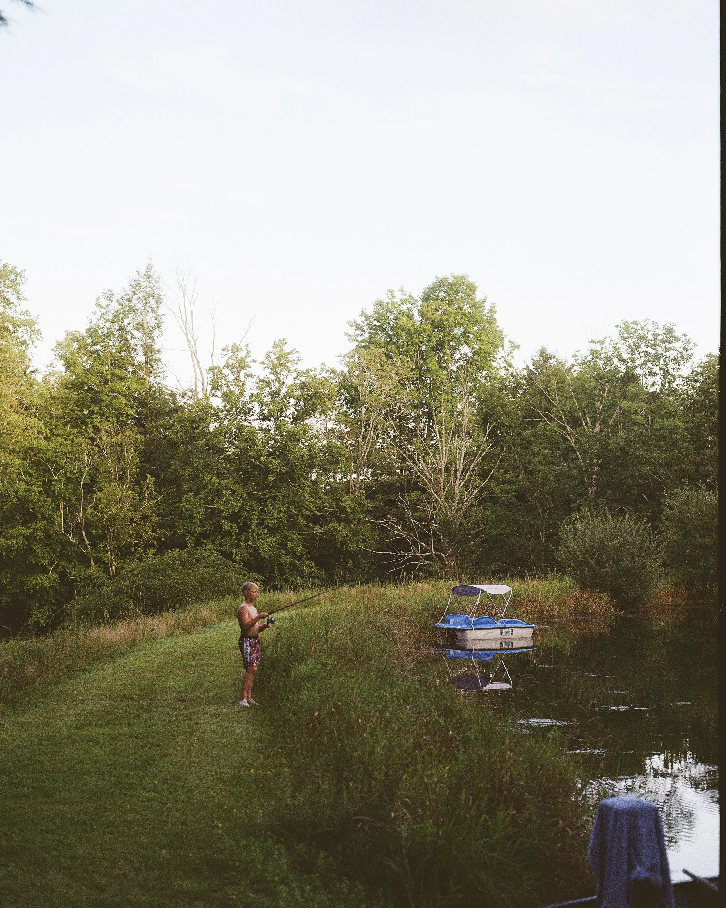 A young boy in swim shorts fishing in a grassy area near a calm body of water, with a small pedal boat docked nearby and lush green trees in the background during daylight.