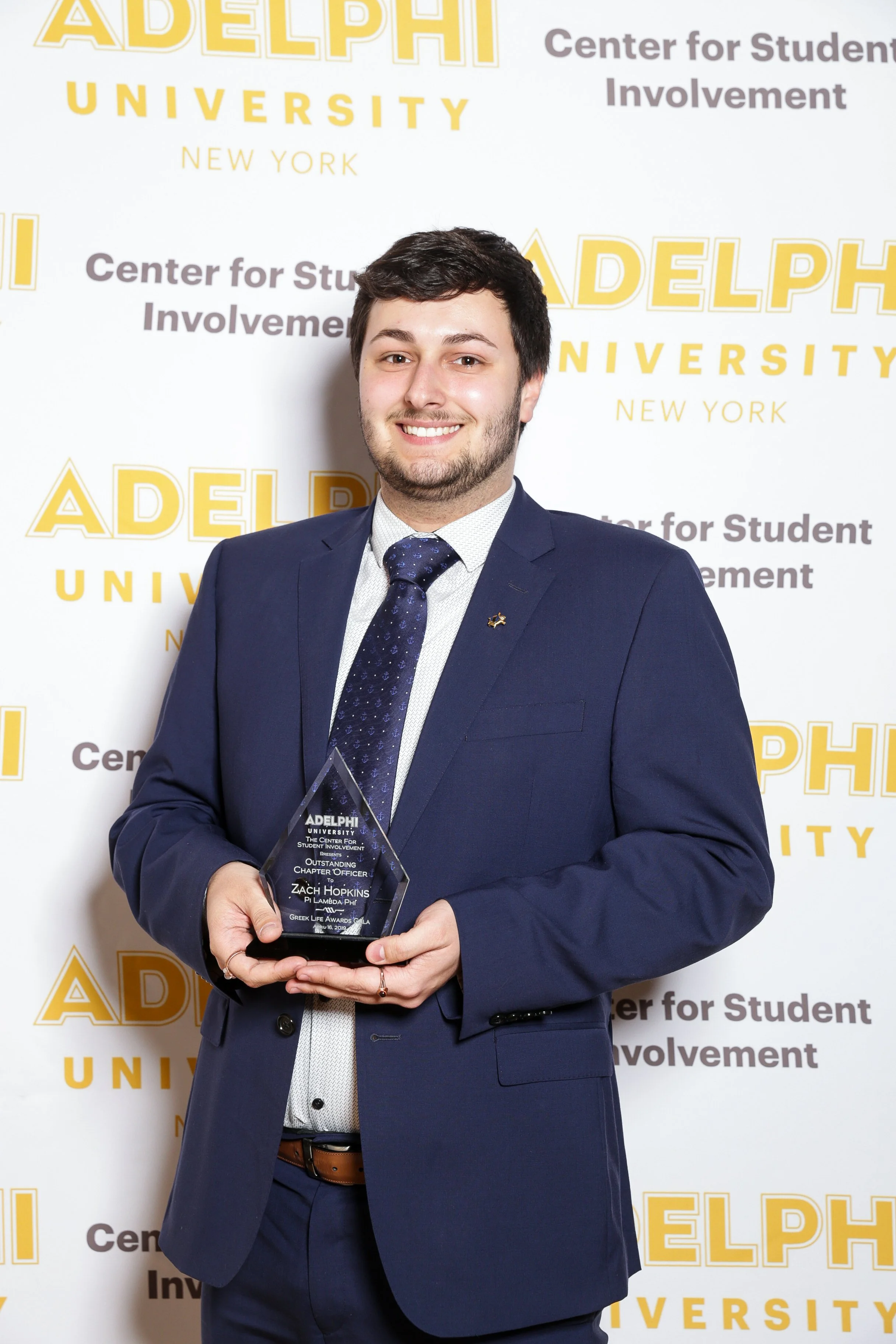 A young man in a navy suit holding an award, standing in front of a backdrop with the words 'Adelphi University' and 'Center for Student Involvement' printed repeatedly.