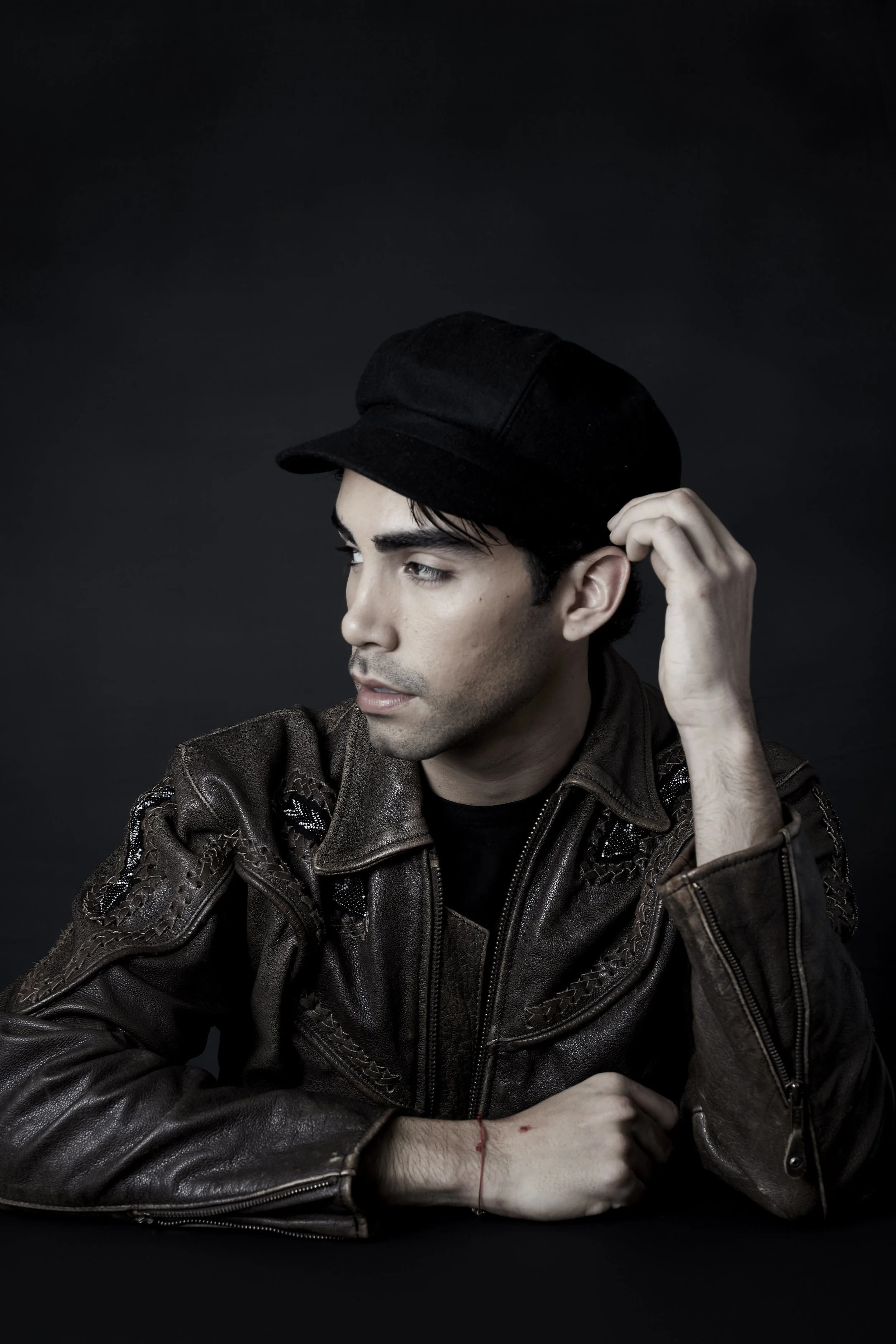 A young man with dark hair and light skin, wearing a black cap and a brown leather jacket with embroidered detailing, posing against a dark background. He looks to the side with a thoughtful expression and has a red string bracelet on his wrist.