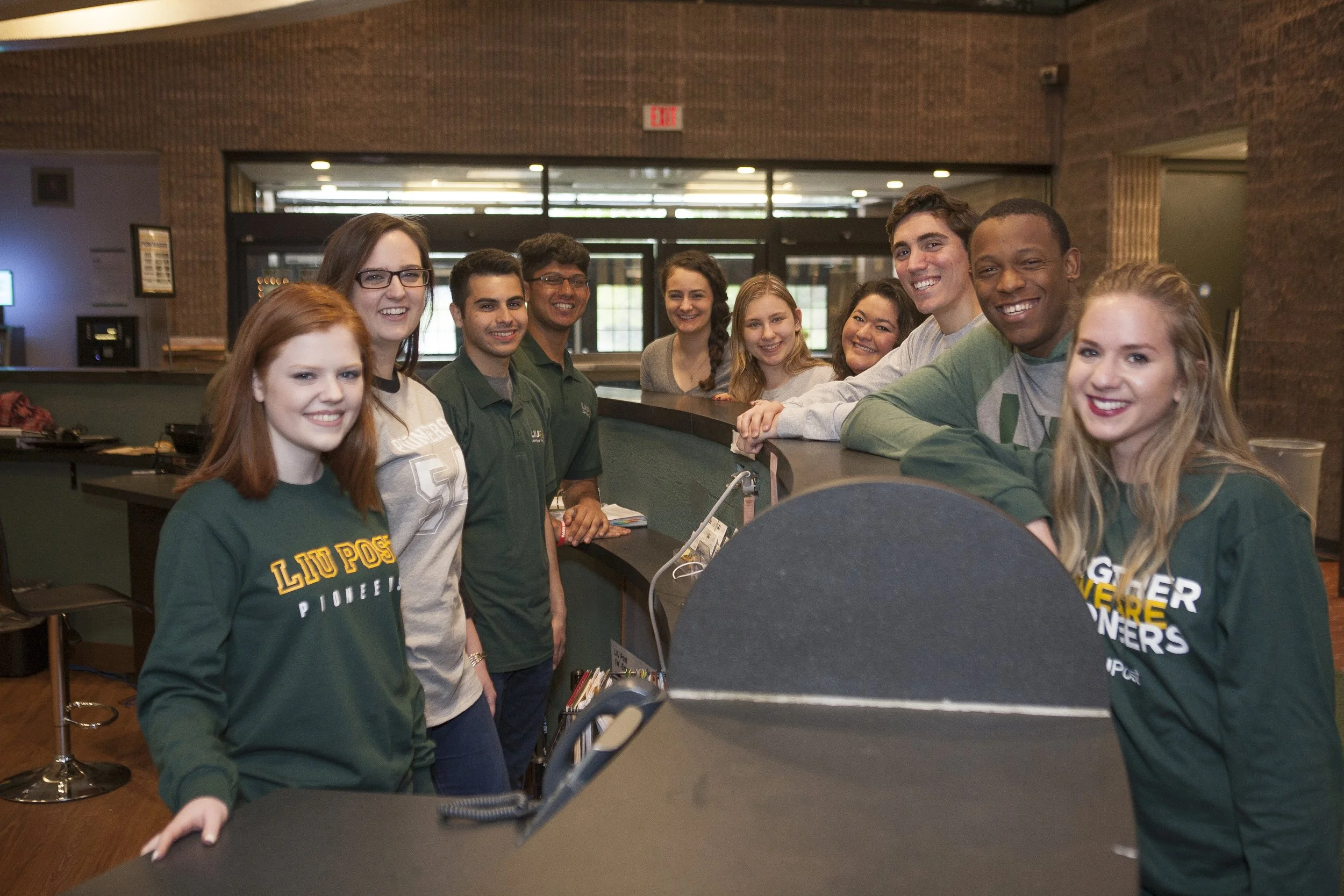 Group of smiling young adults standing at a reception desk in a lobby or restaurant.