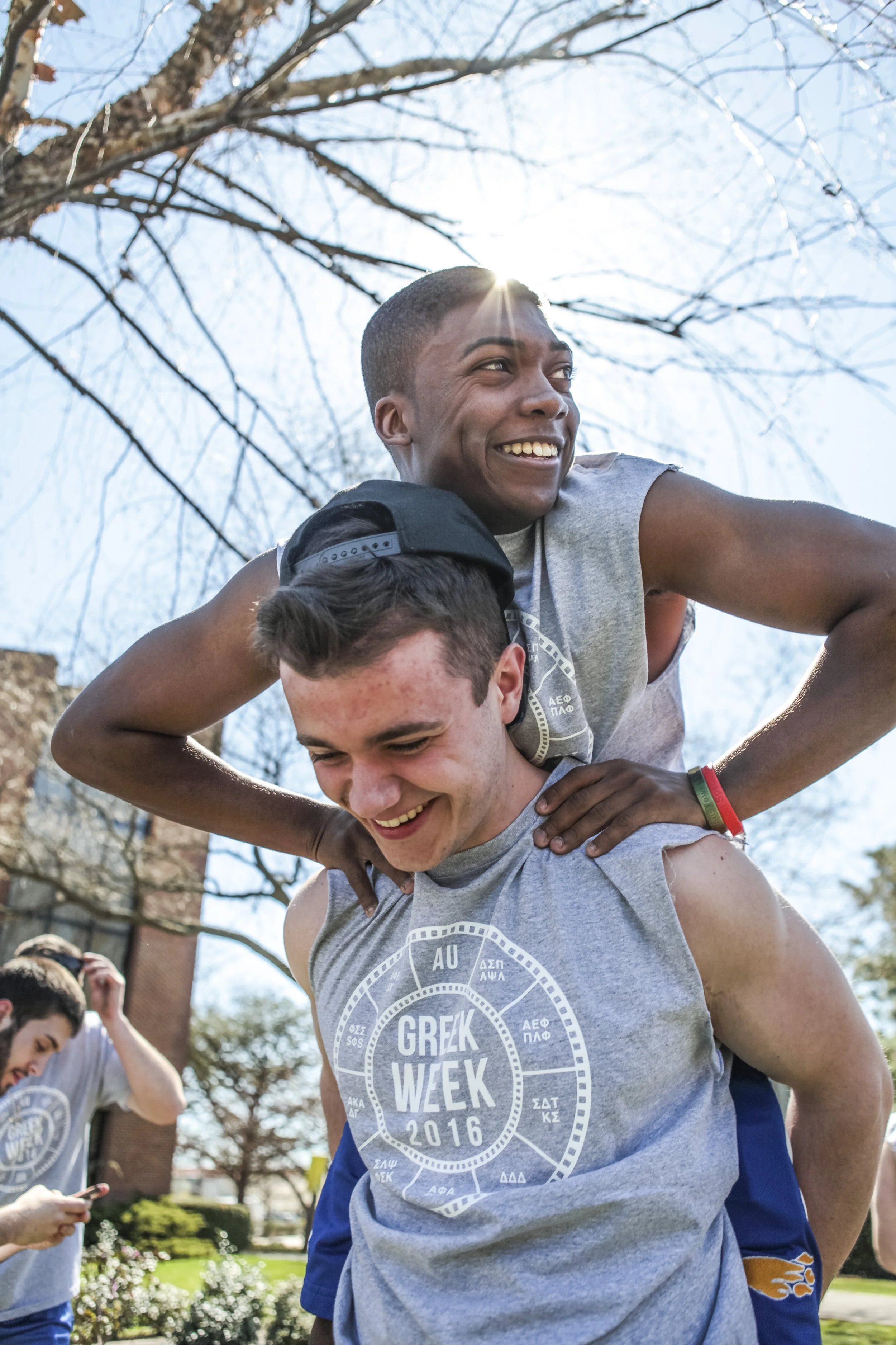 Two young men participating in a Greek Week event outdoors, with one giving the other a piggyback ride, both smiling, on a sunny day with bare trees and a brick building in the background.