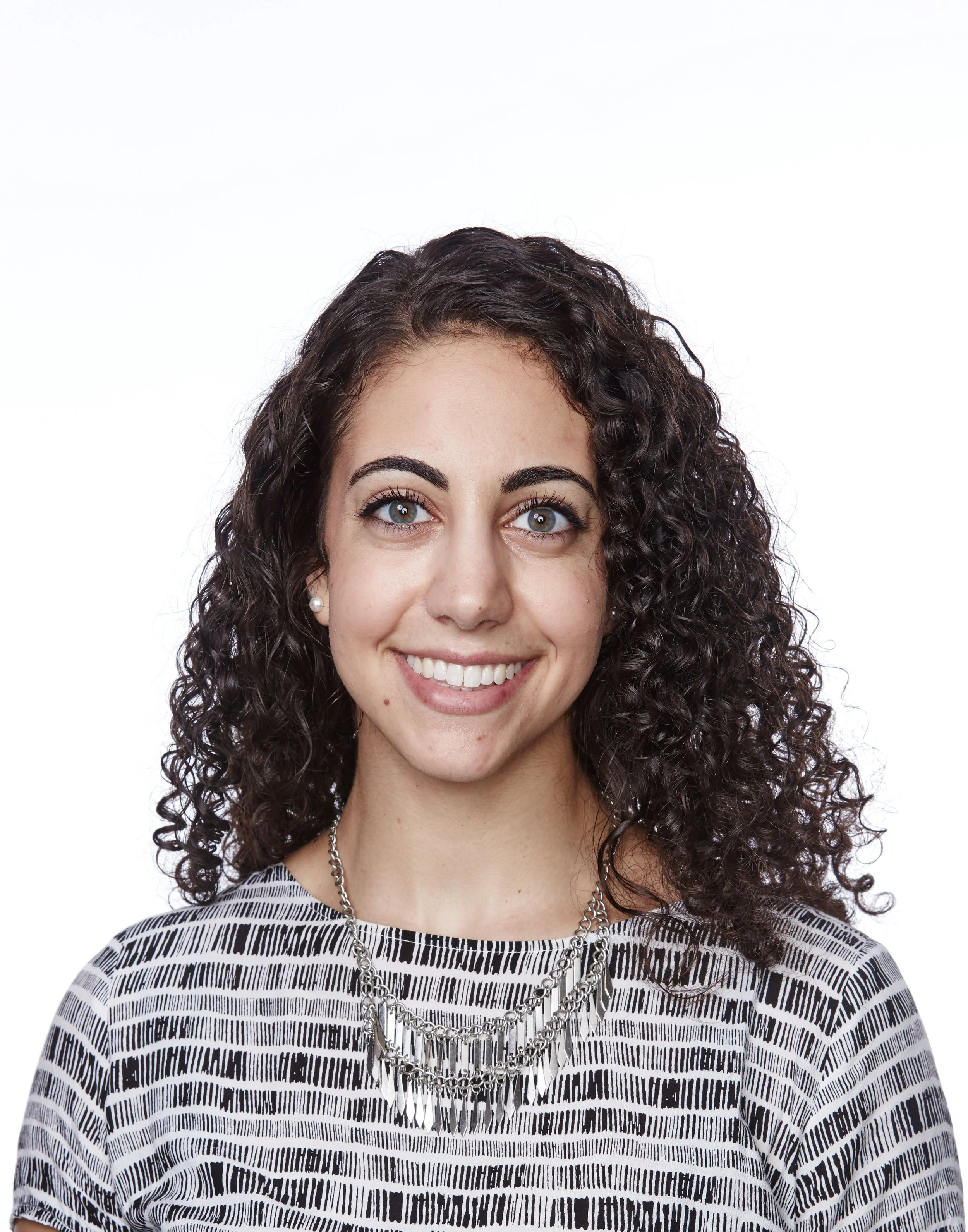 Portrait of a young woman with curly dark hair and blue eyes, smiling, wearing a black and white striped top and layered silver necklaces against a white background.