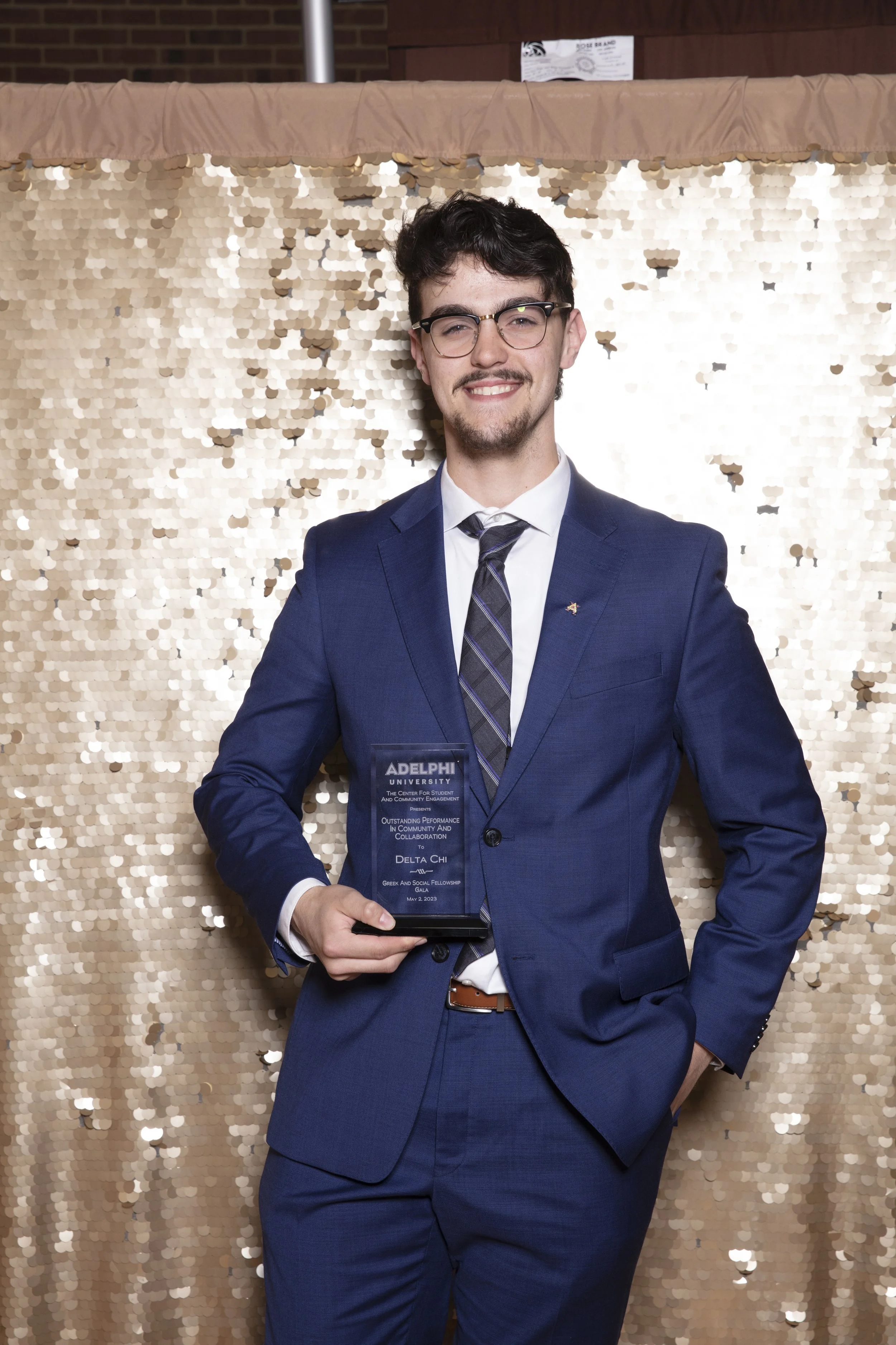 Young man in a blue suit holding a plaque at an awards event, with a gold sequin backdrop.