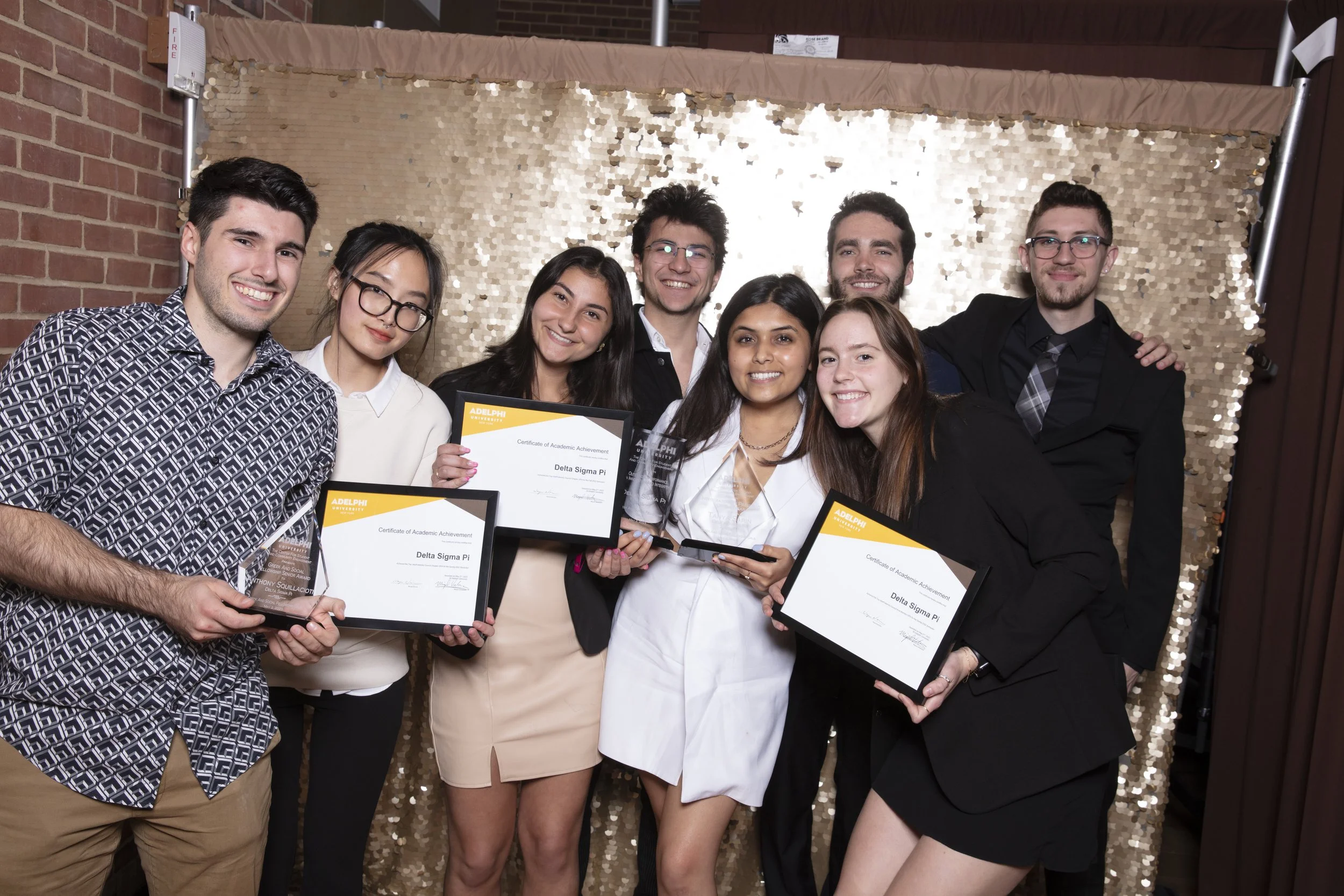 Group of eight young adults at an awards ceremony, holding certificates and trophies, smiling, standing in front of a gold sequin backdrop.