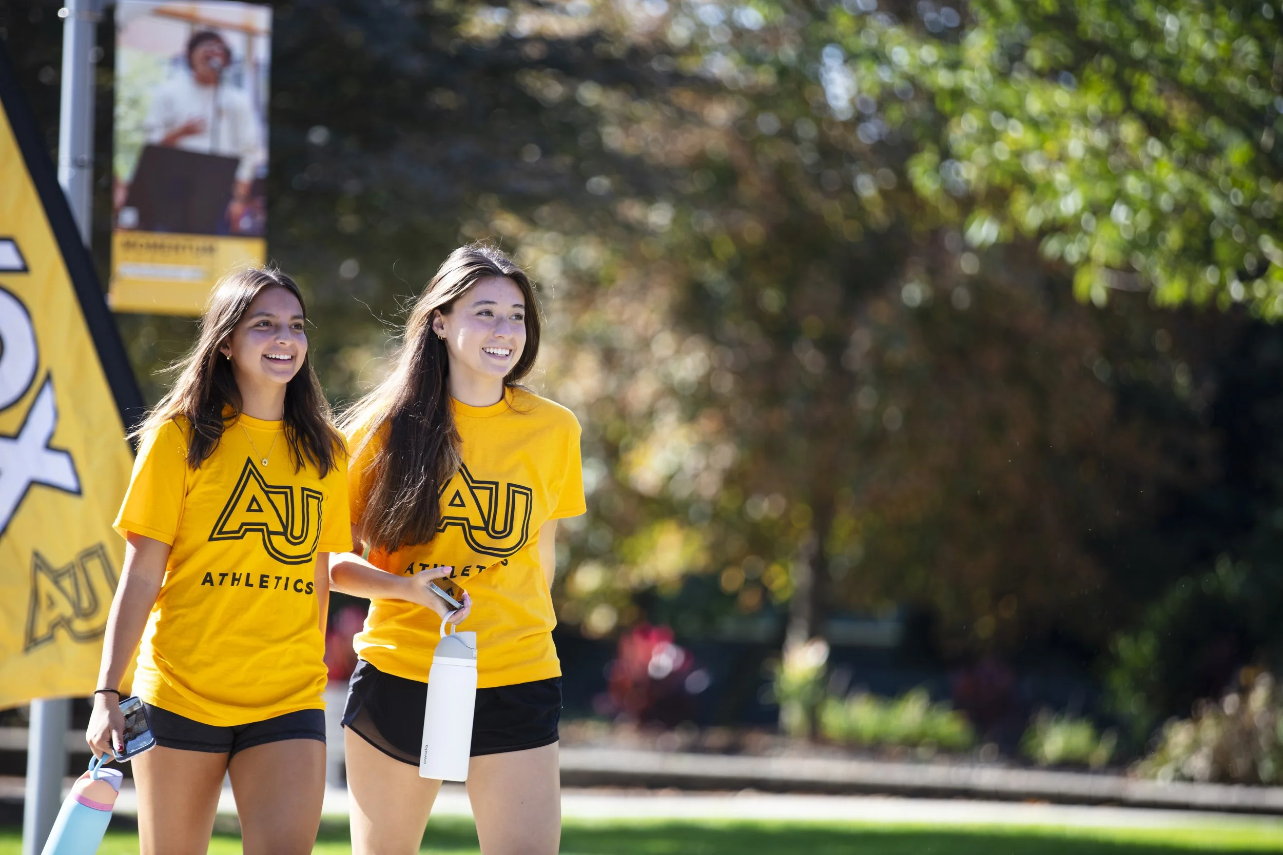 Two young women wearing yellow Auburn University Athletics t-shirts, walking outdoors on a sunny day, smiling and holding water bottles.