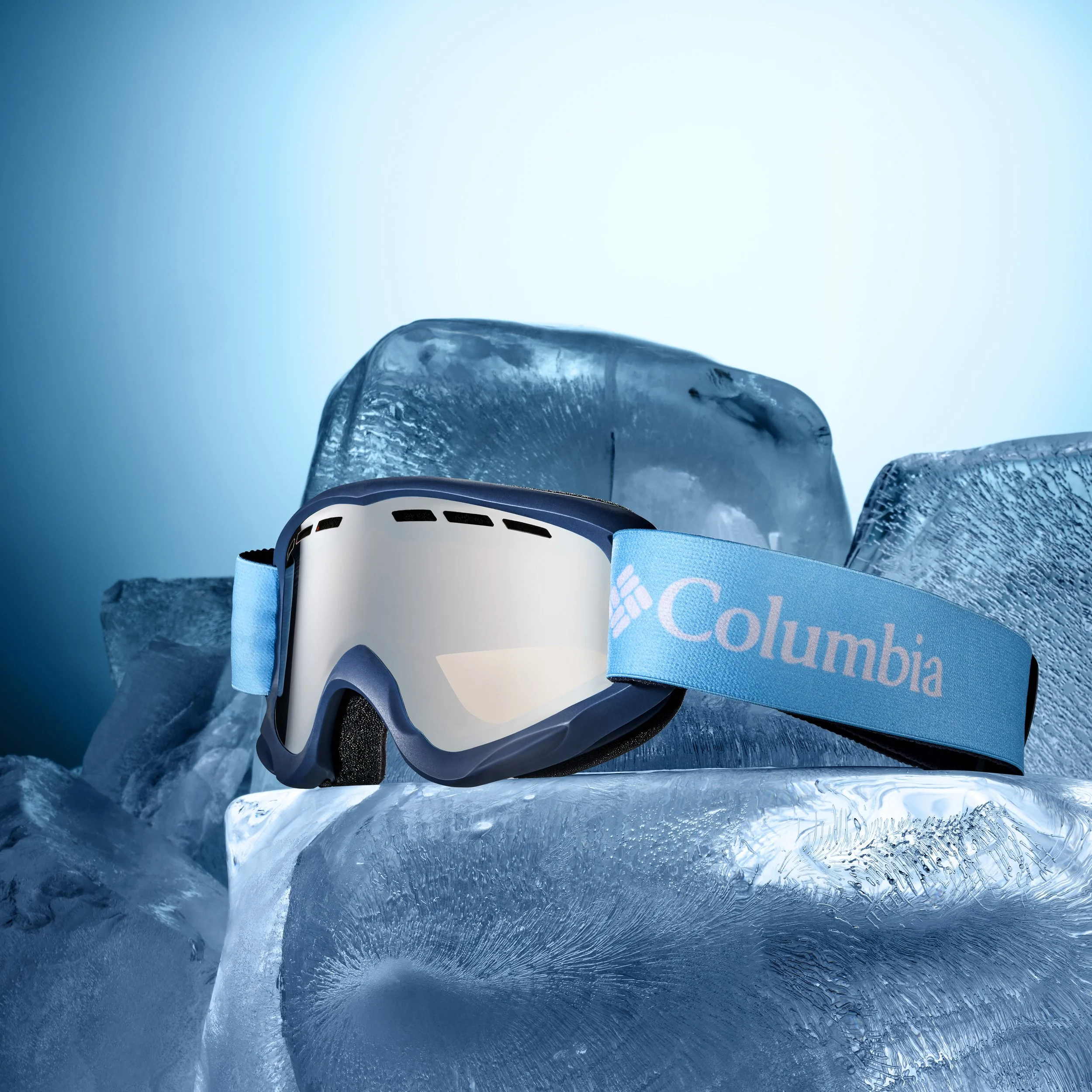 A Columbia ski goggle with a mirrored lens, resting on a block of ice with larger ice blocks in the background, under cool blue lighting.