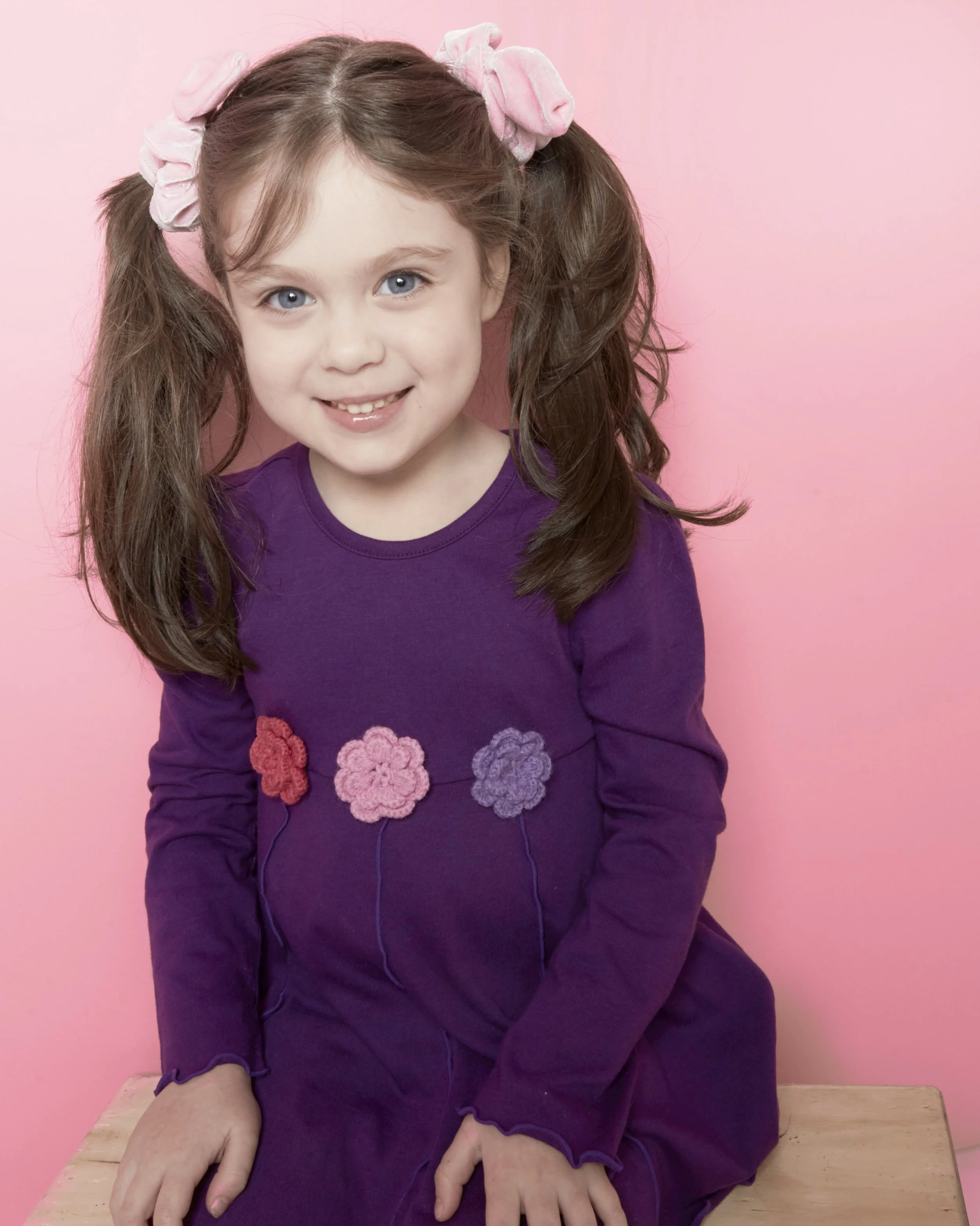 A young girl with long brown hair in pigtails, wearing pink scrunchies, a purple dress with pink and purple floral decorations, sitting on a wooden surface with a pink background, smiling at the camera.