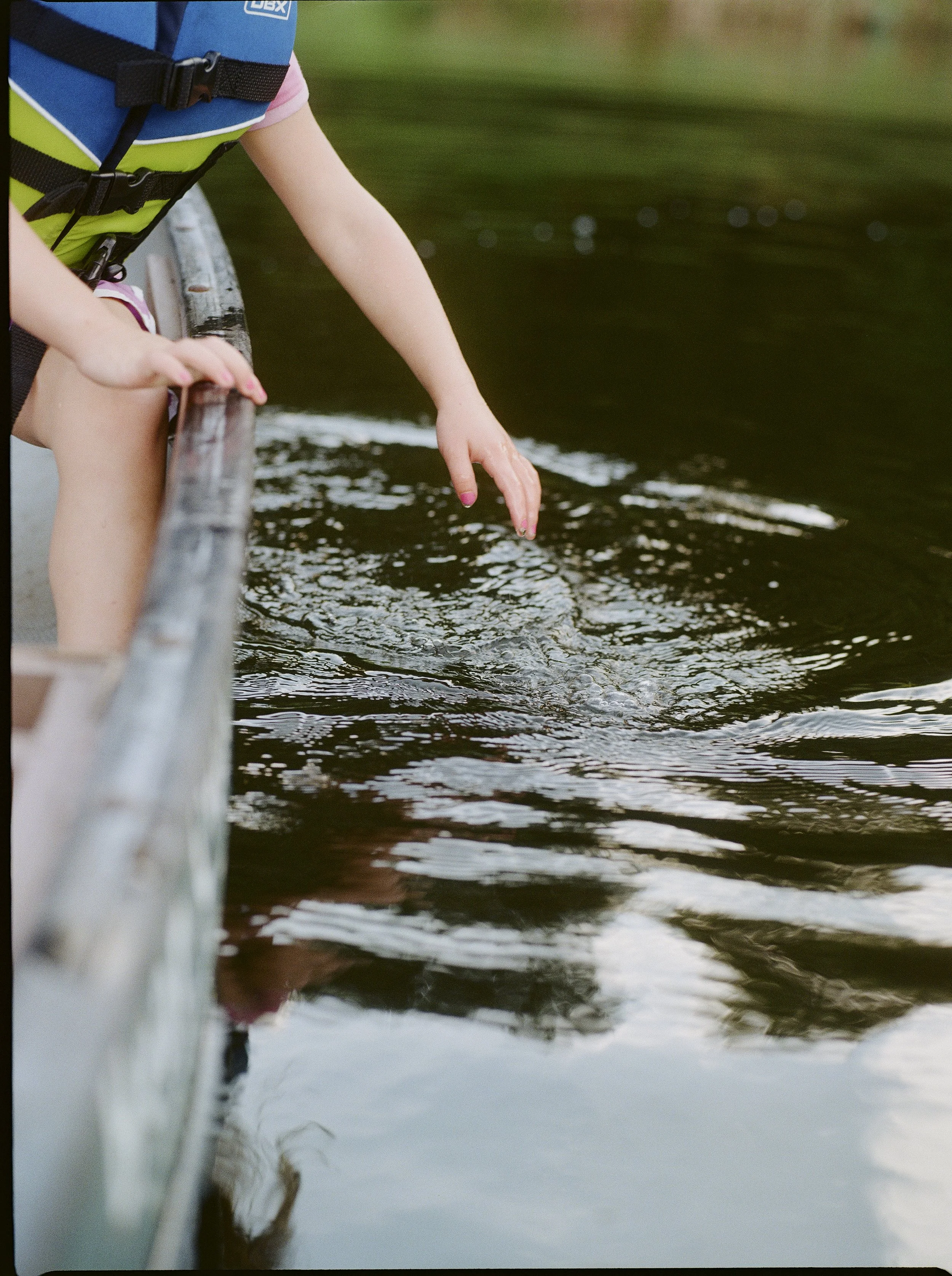 Child leaning over a wooden railing, touching the water in a river or lake during daytime, wearing a life jacket.