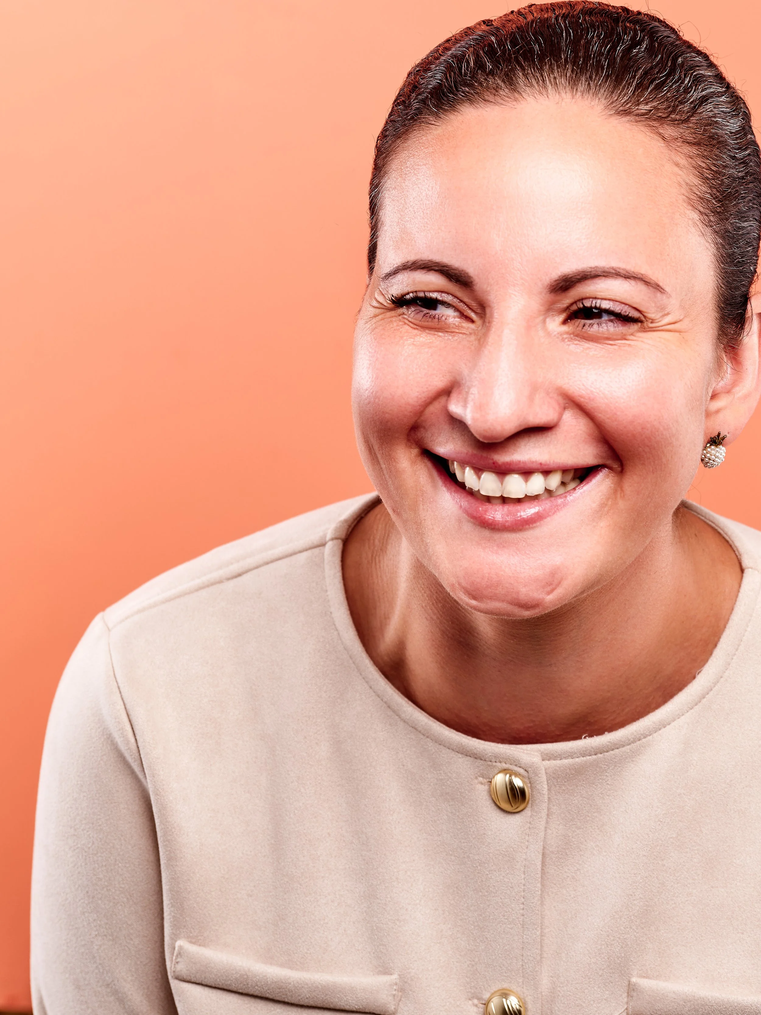 Close-up of a woman smiling, wearing pearl earrings and a beige blazer with gold buttons, against a peach-colored background.