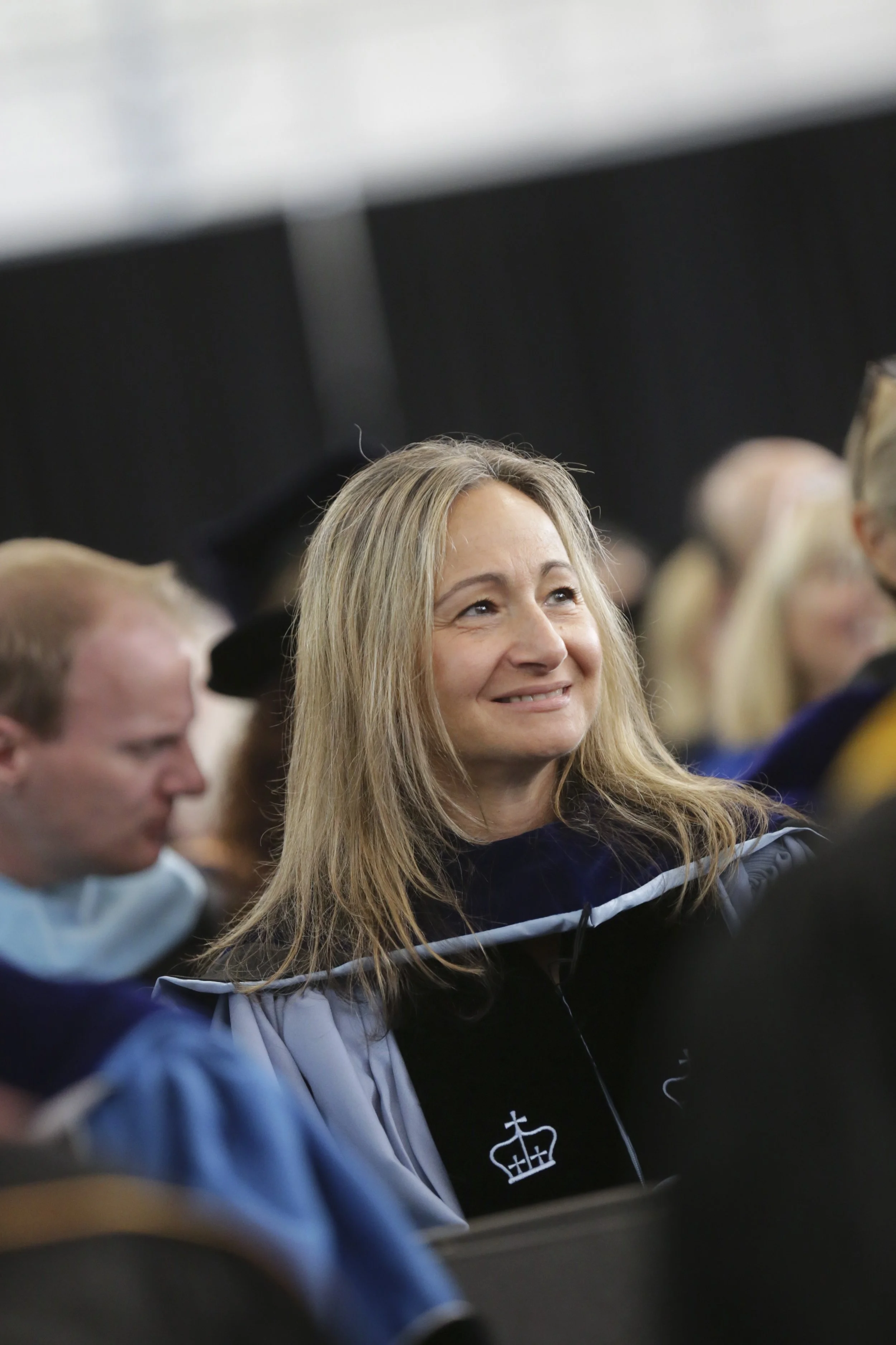 A woman with blonde hair smiling during a graduation ceremony, surrounded by other graduates in academic regalia.