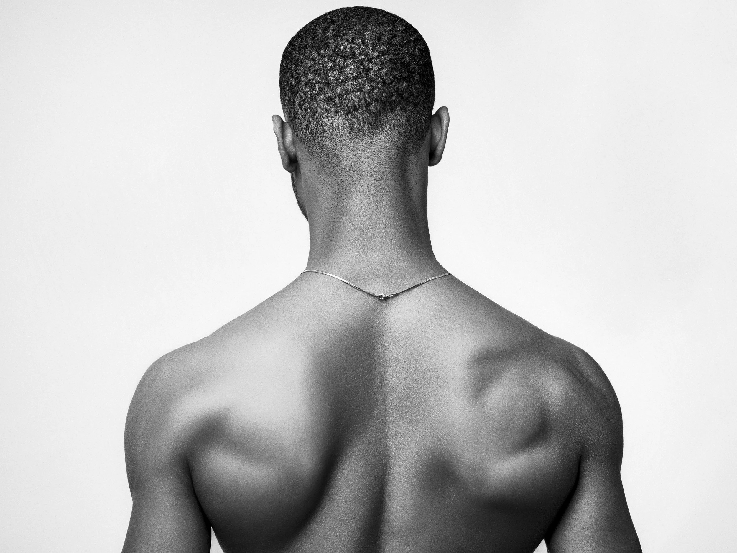 Back view of a shirtless man with short textured hair, wearing a thin chain necklace, against a plain background.