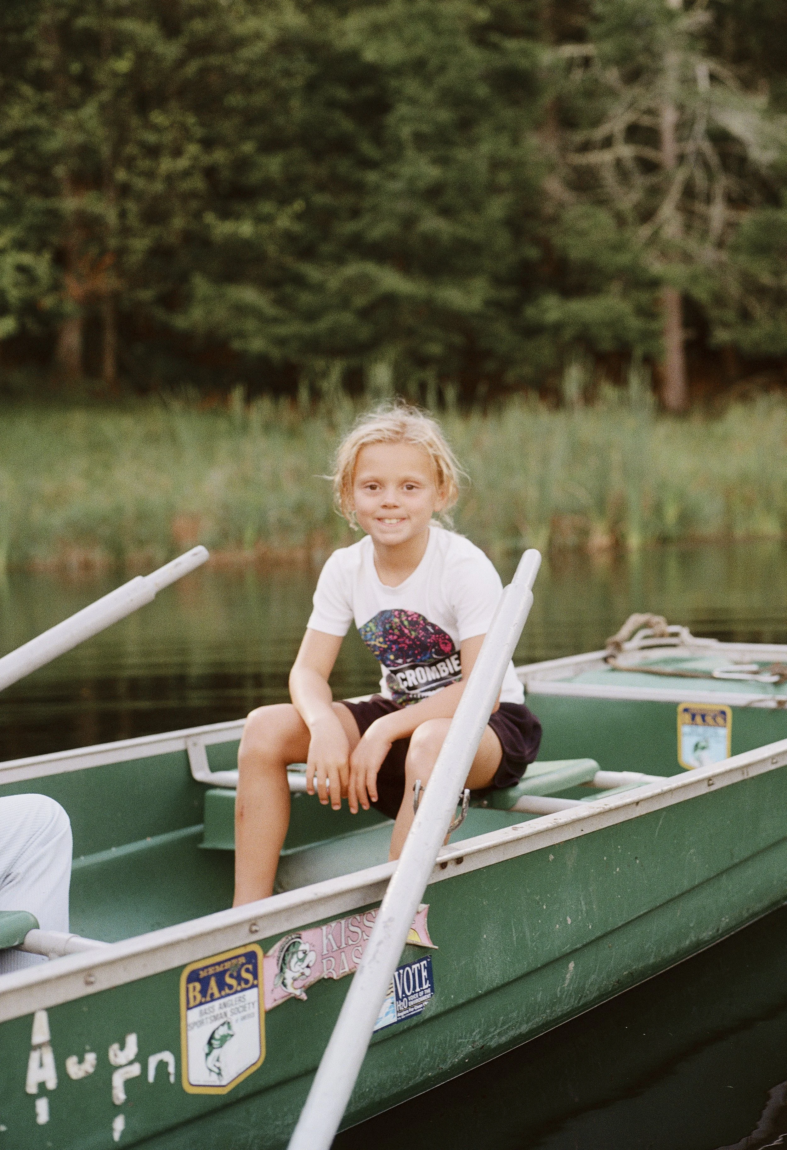 A young boy with blond hair sitting in a green boat on a lake, smiling, with dense trees in the background.