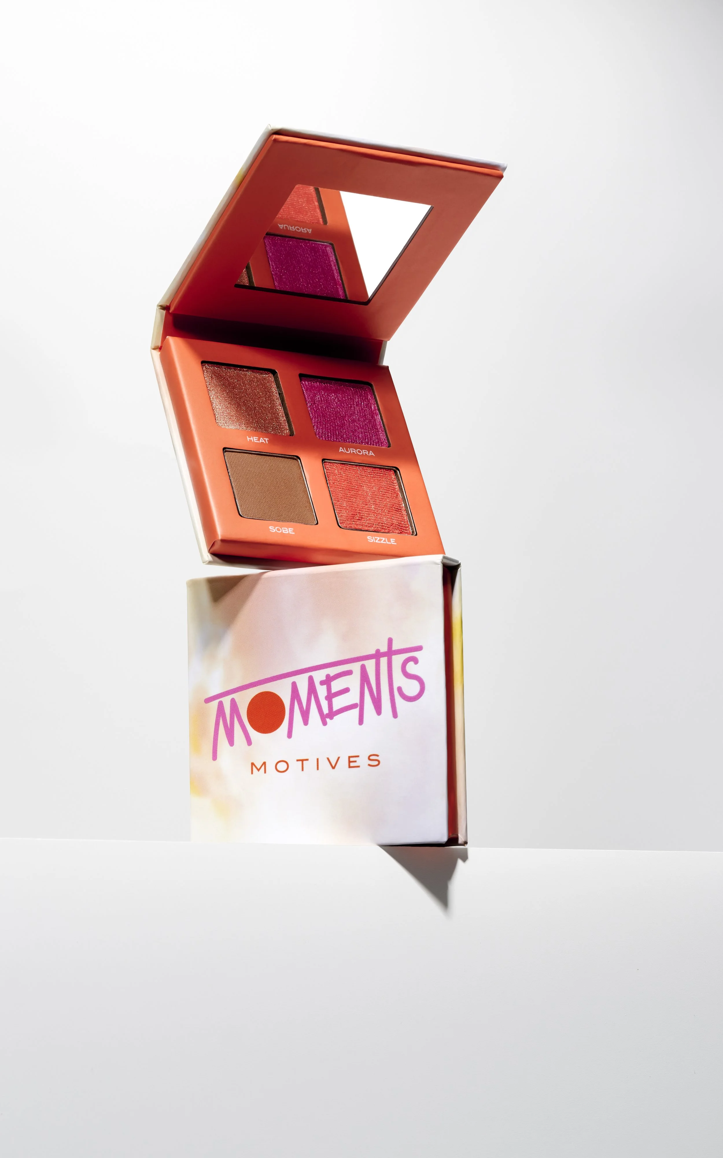 Open makeup palette with four eyeshadow shades above a box labeled 'Moments Motives', on a white surface.