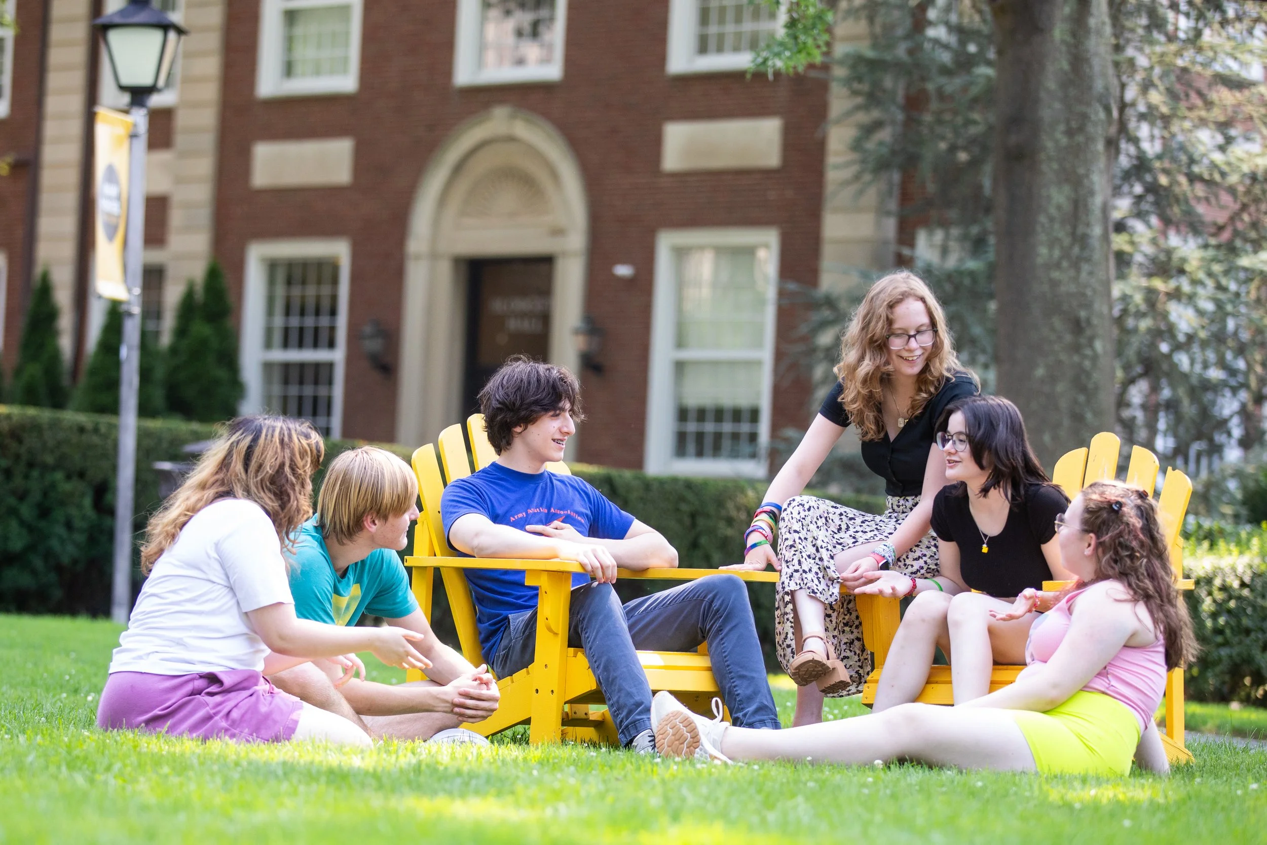 Group of young friends sitting and talking on grass near yellow Adirondack chairs outdoors.