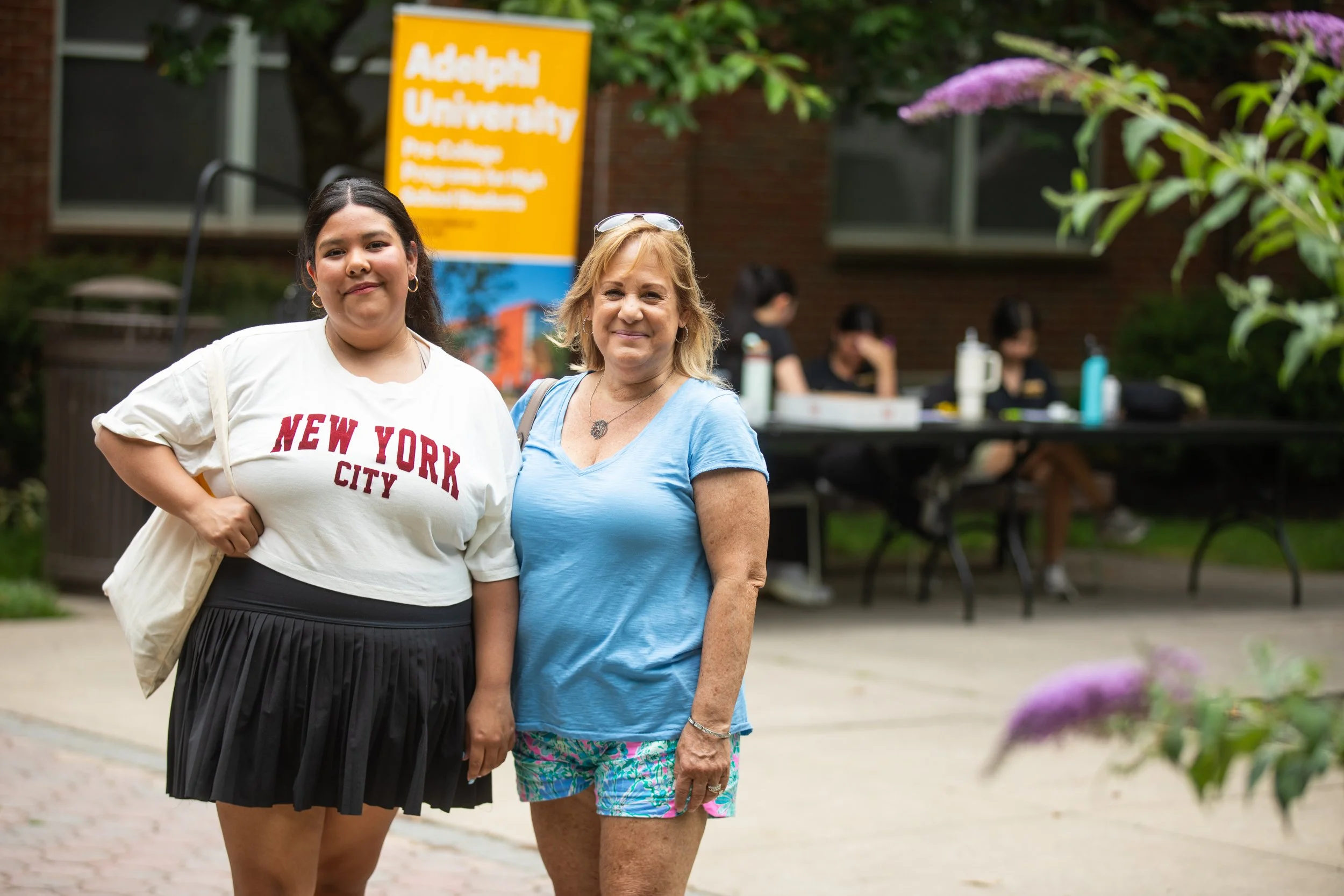 Two women standing outdoors in front of a yellow Adelphi University sign; the woman on the left wears a white t-shirt with 'New York City' printed and a black pleated skirt, the woman on the right wears a light blue t-shirt and colorful shorts.