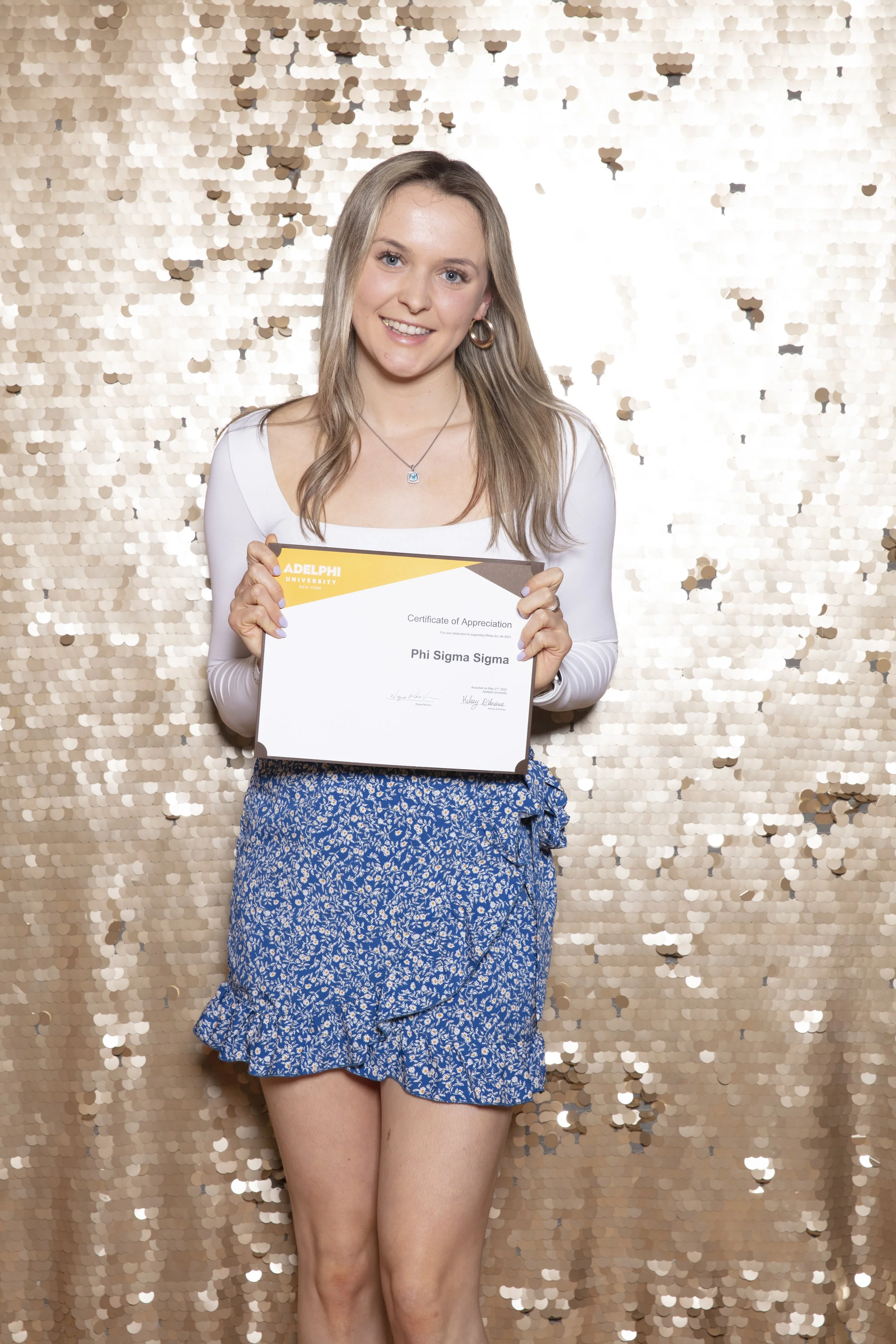 A young woman with long blonde hair, blue eyes, and hoop earrings holding a diploma, standing in front of a gold sequin backdrop.