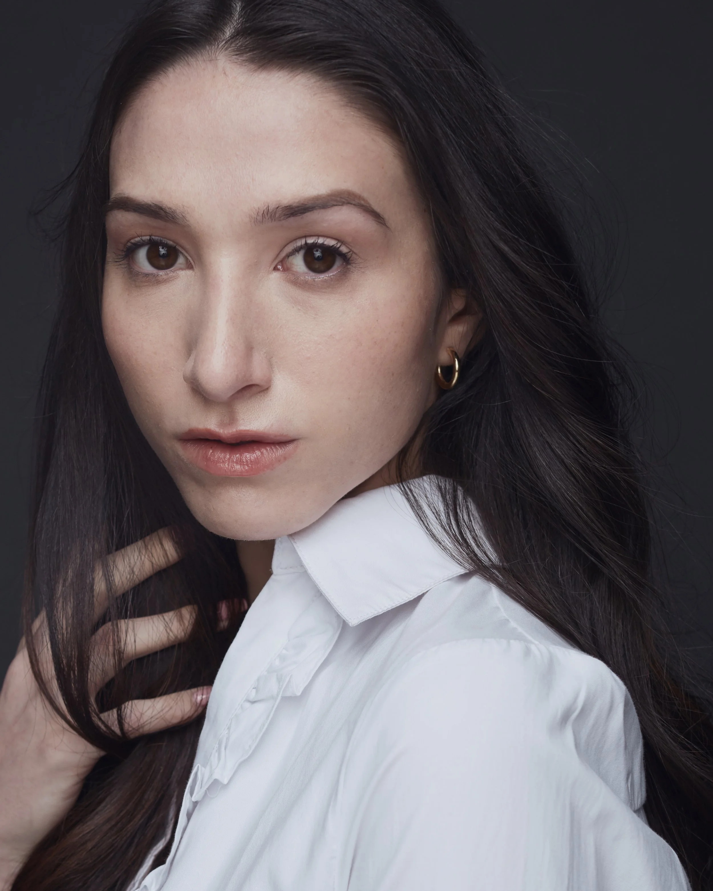 A woman with long dark hair wearing a white shirt, looking at the camera with a neutral expression against a dark background.