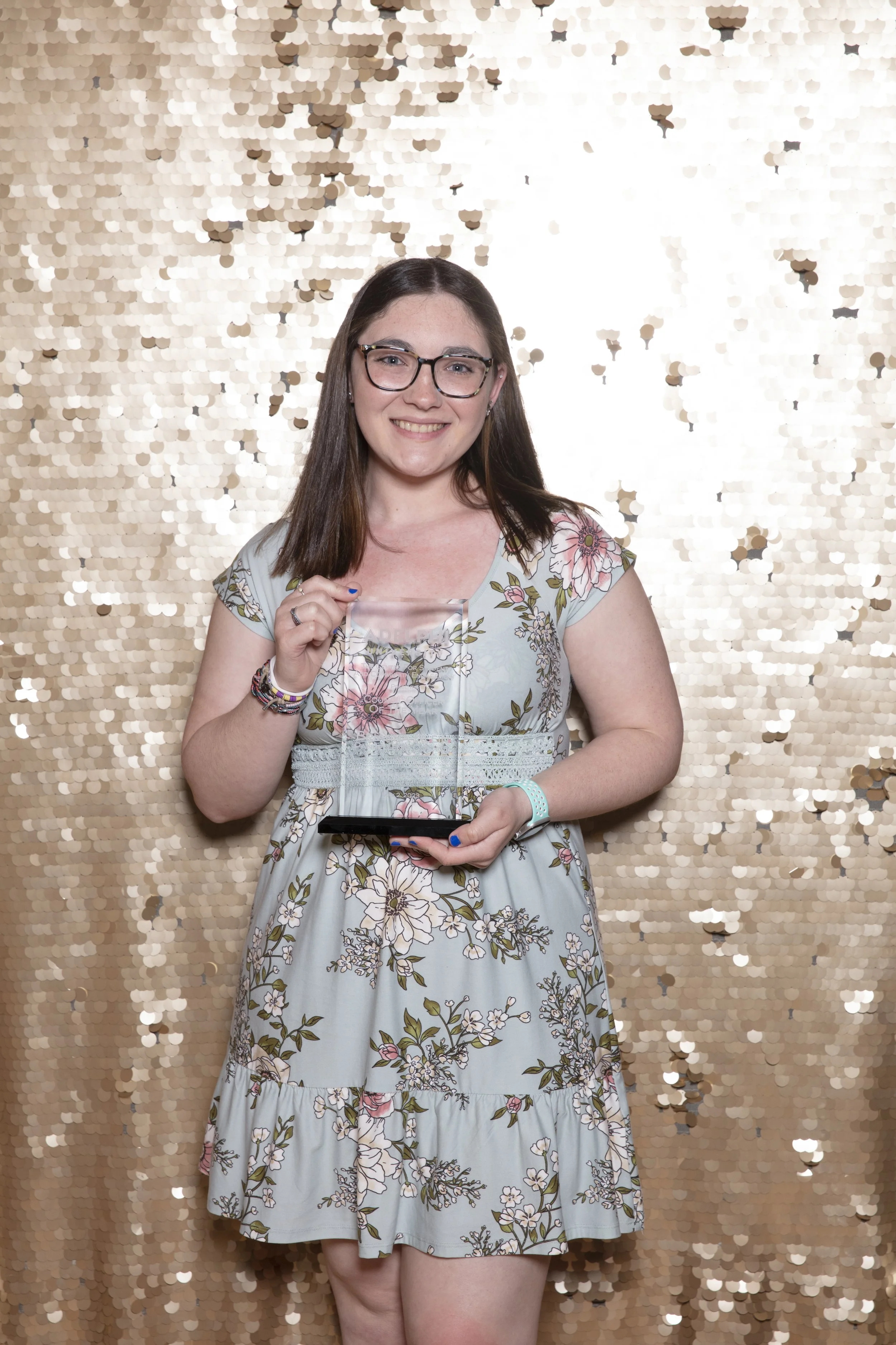 A young woman with glasses and straight brown hair smiling, standing in front of a gold sequin backdrop, holding a clear award and a phone.