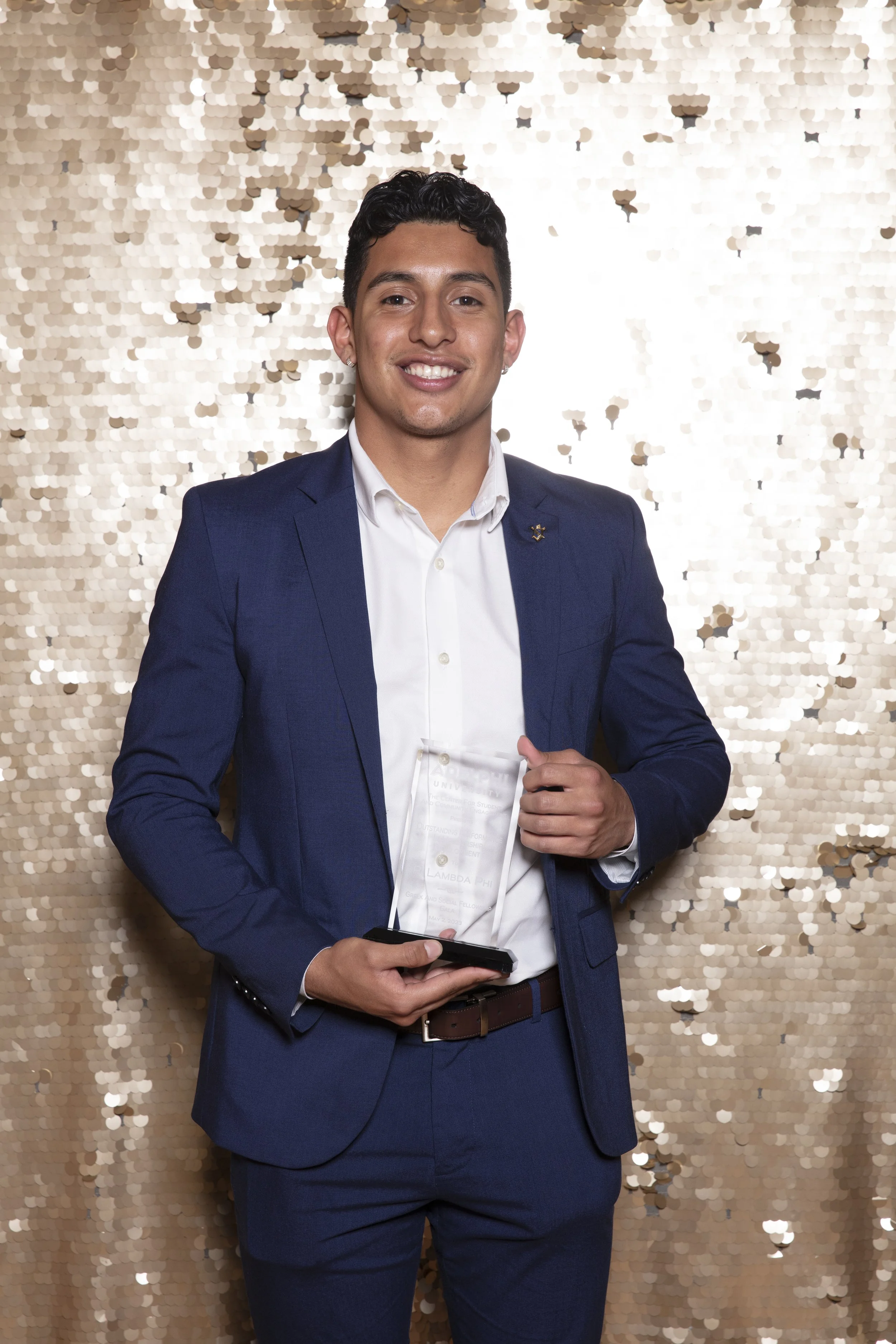 Young man in a blue suit holding an award, standing against a gold sequin backdrop, smiling.