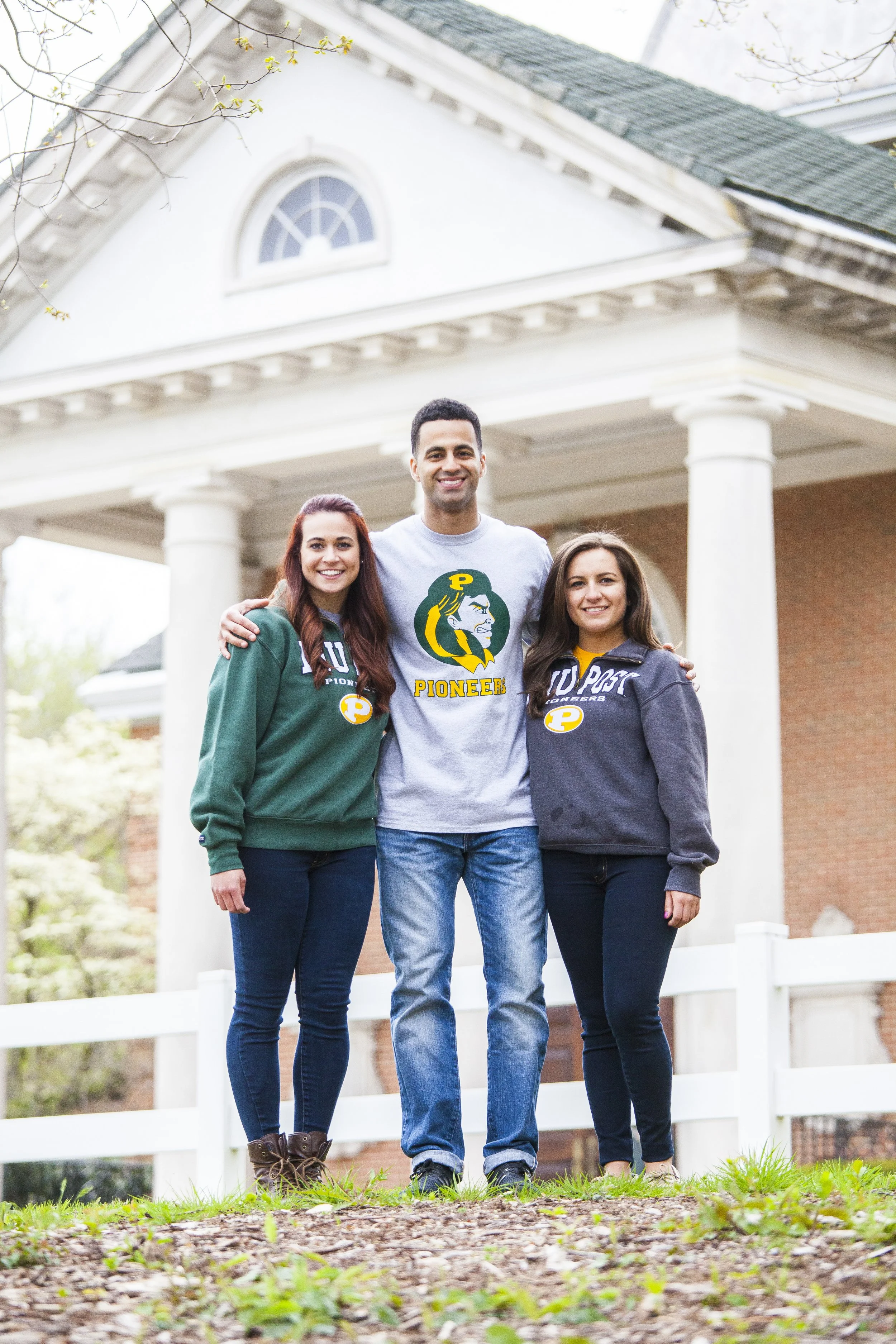 Three young adults standing outdoors in front of a white house or building with columns, smiling, with two women wearing University of Portland sweatshirts and a man wearing a Pioneers t-shirt.