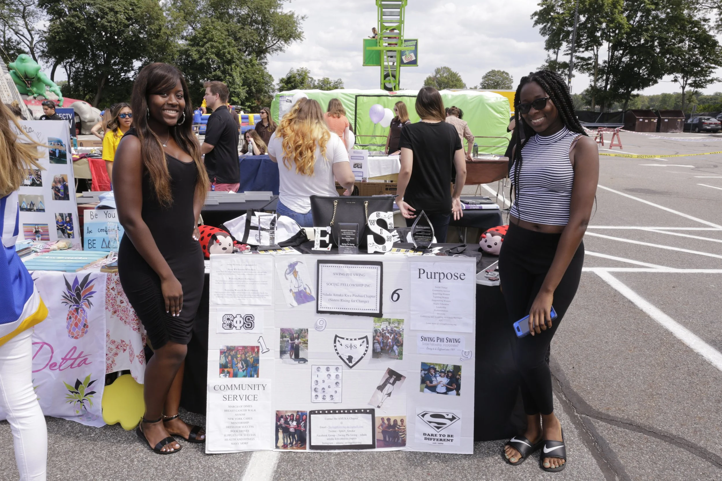 Two women standing beside a community service booth at an outdoor event, with other booths and people in the background.