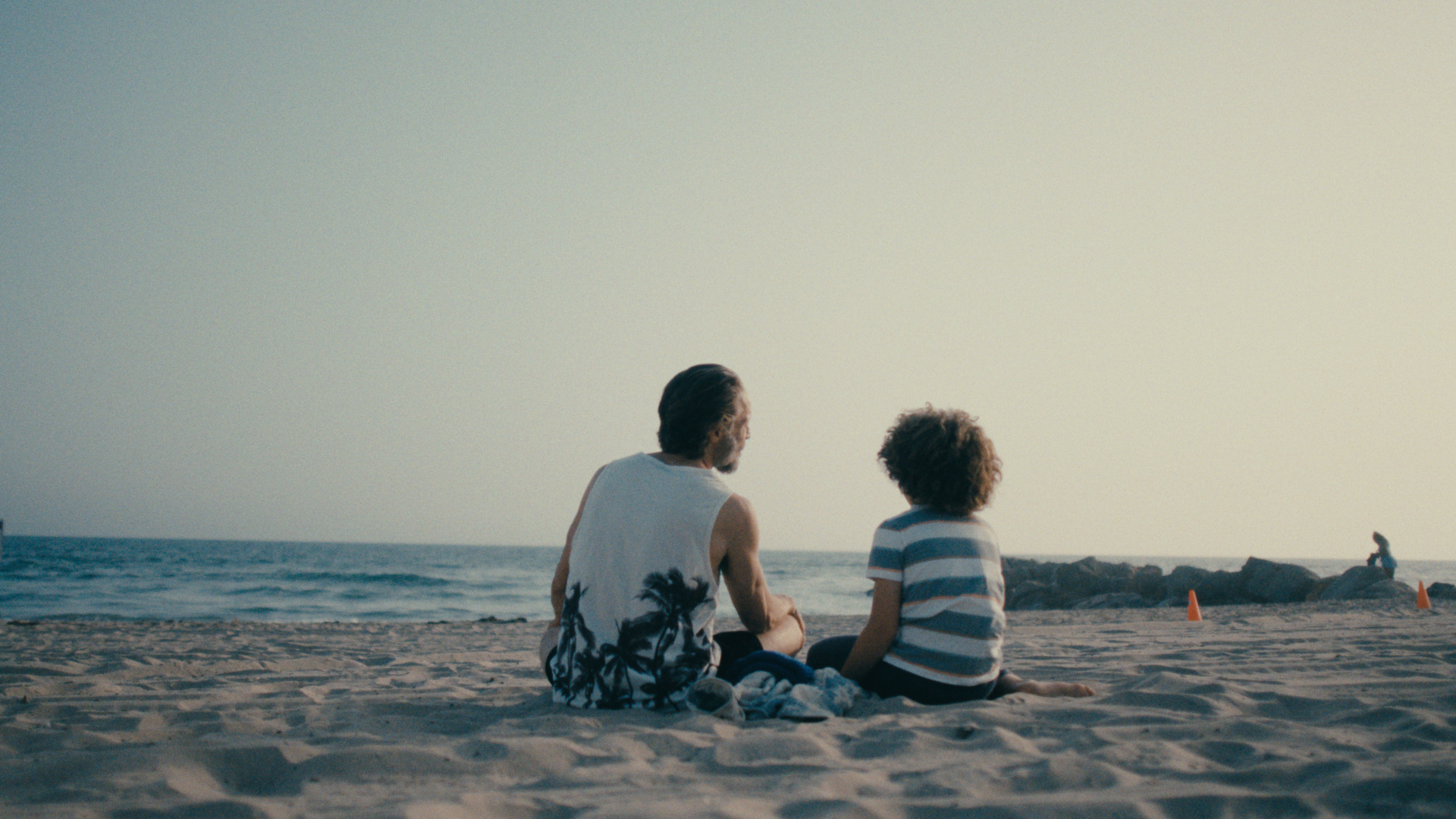 A man and a child sitting on the sandy beach looking at the ocean, with orange cones and rocks in the background.