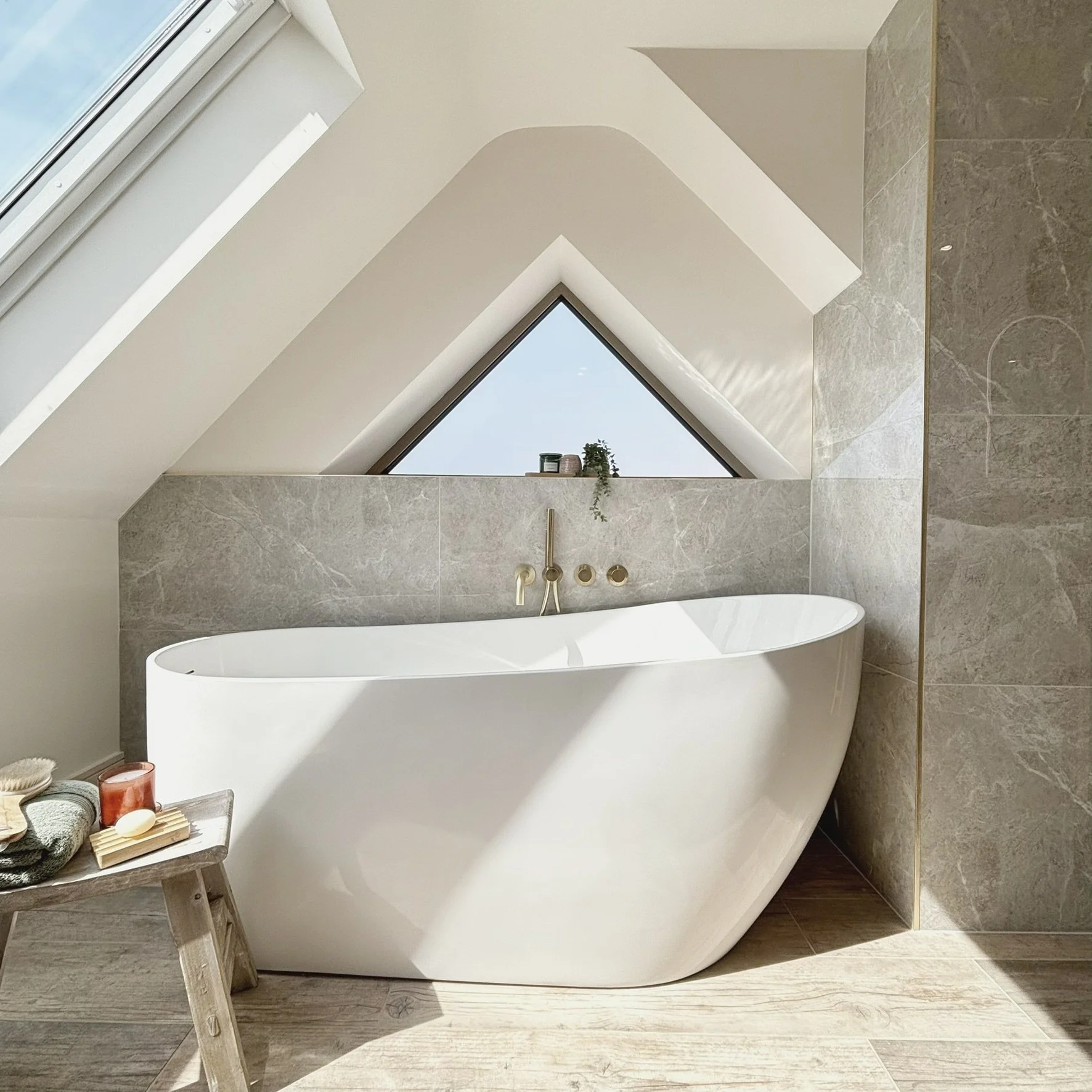 A modern bathroom featuring a freestanding white bathtub, beige tiled walls, and a triangular window with decorative plants on the window ledge.