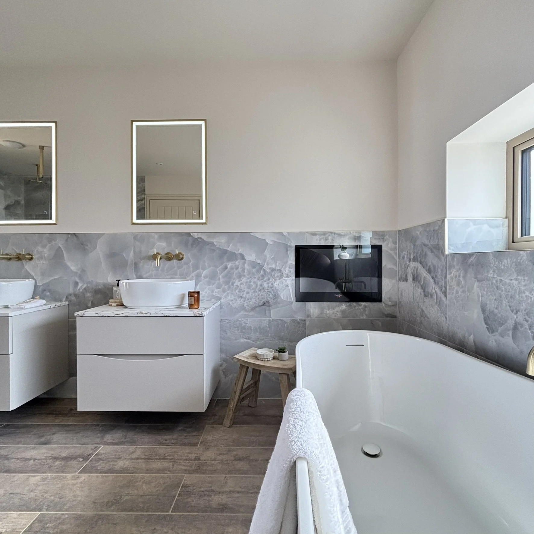 Modern bathroom with wall hung vanity, vessel sinks, brushed brass brassware, blue/gray marble wall tiles, a freestanding bathtub looking onto a wall mounted TV.