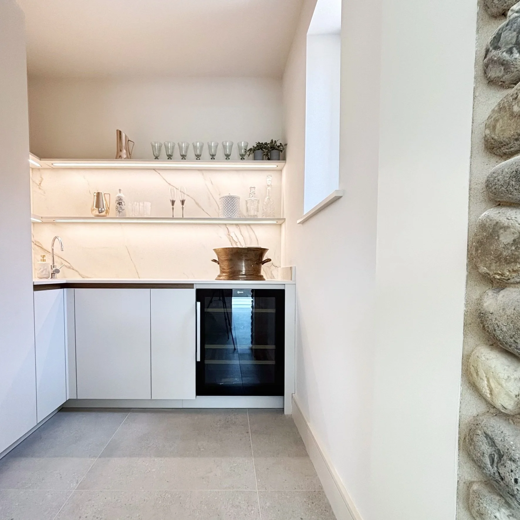 A modern pantry/bar area with a beach grey cabinets and a built-in wine fridge, shelves with routered lighting underneath.  Ceramic splashback up the wall to match the worktop.