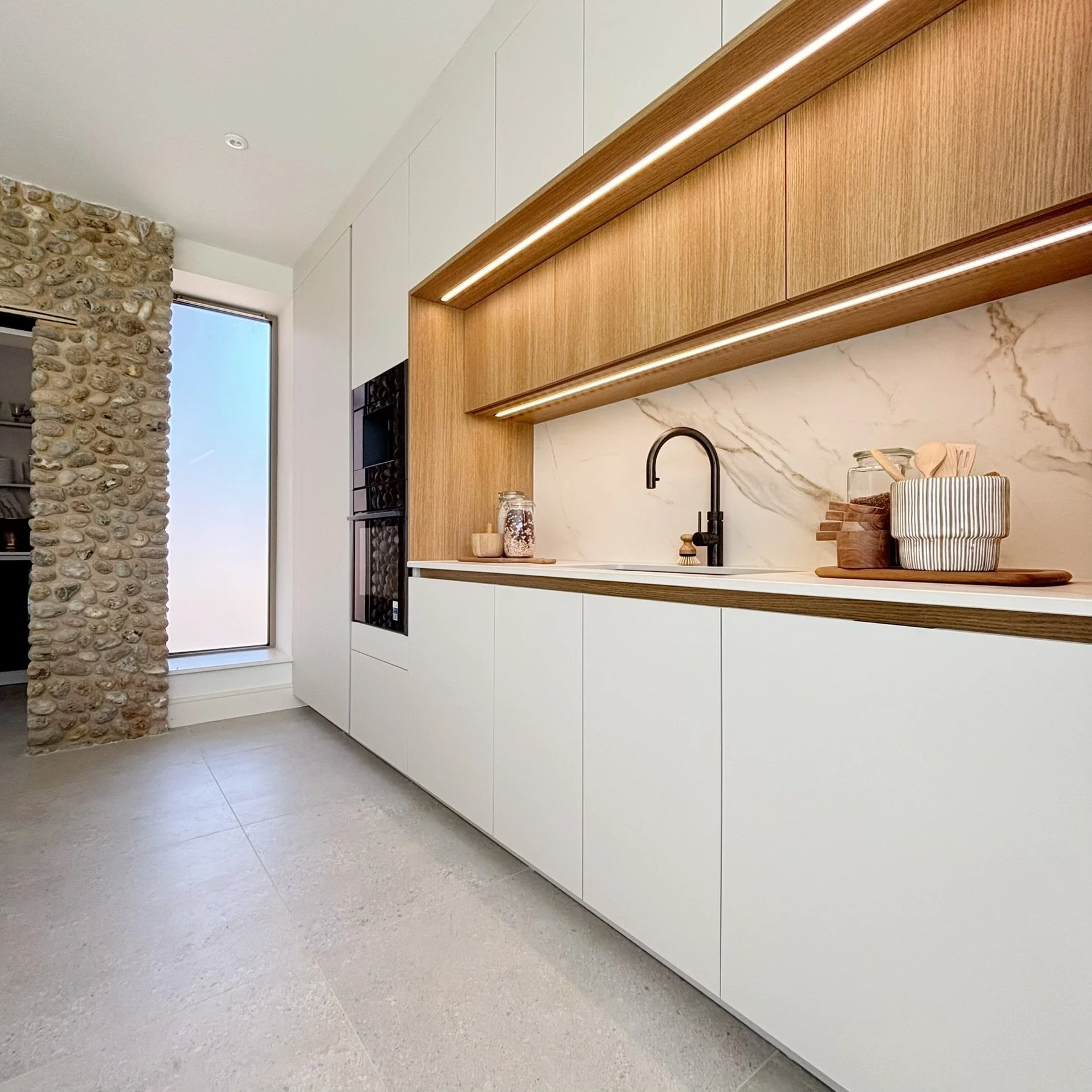 Modern kitchen with white cabinets, wooden open shelves with integrated lighting, a marble backsplash, and a black faucet. A stone wall on the left and a large window letting in natural light.