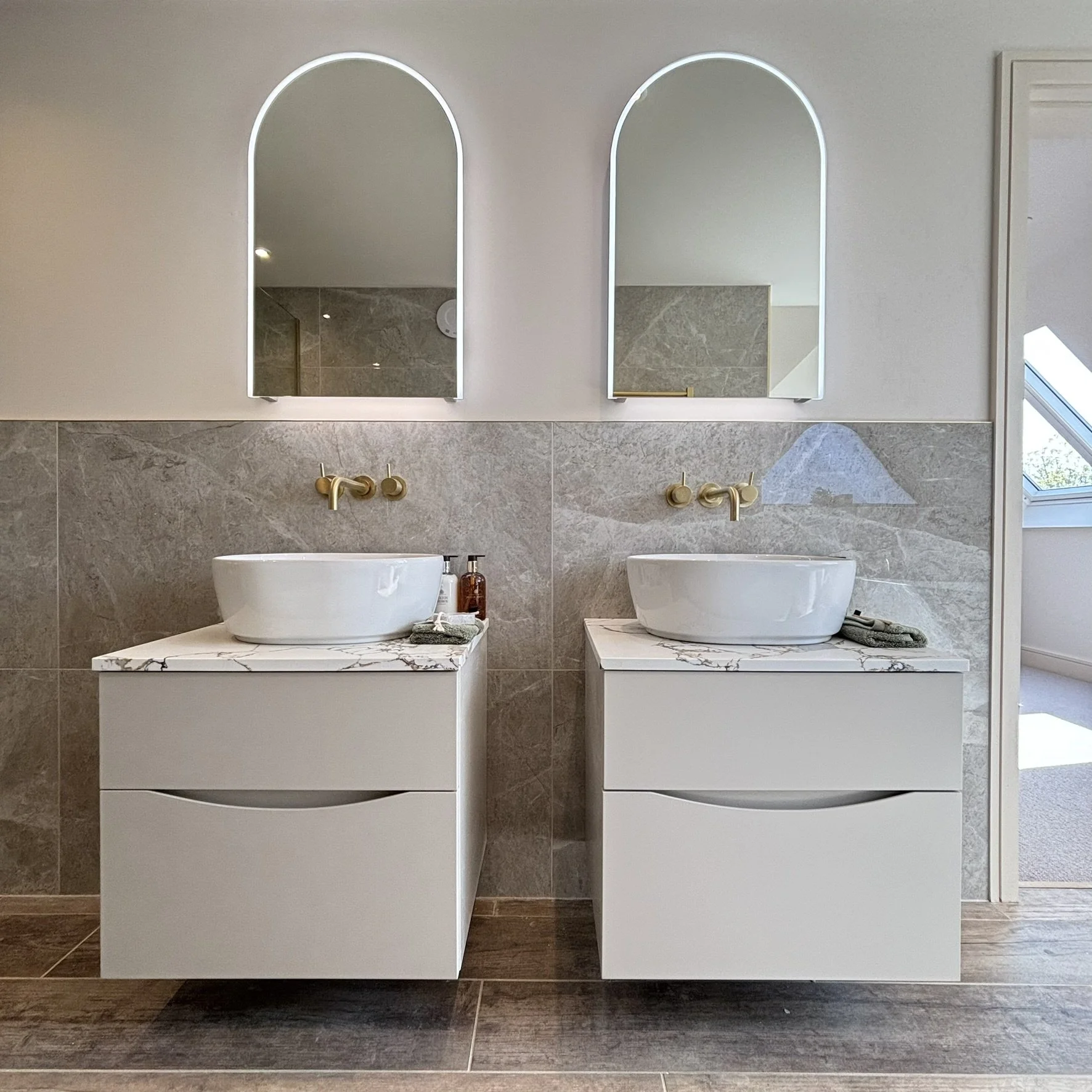 Modern bathroom with two wall hung vanity units, wall-mounted brushed brass taps, two mirrors with LED lighting.