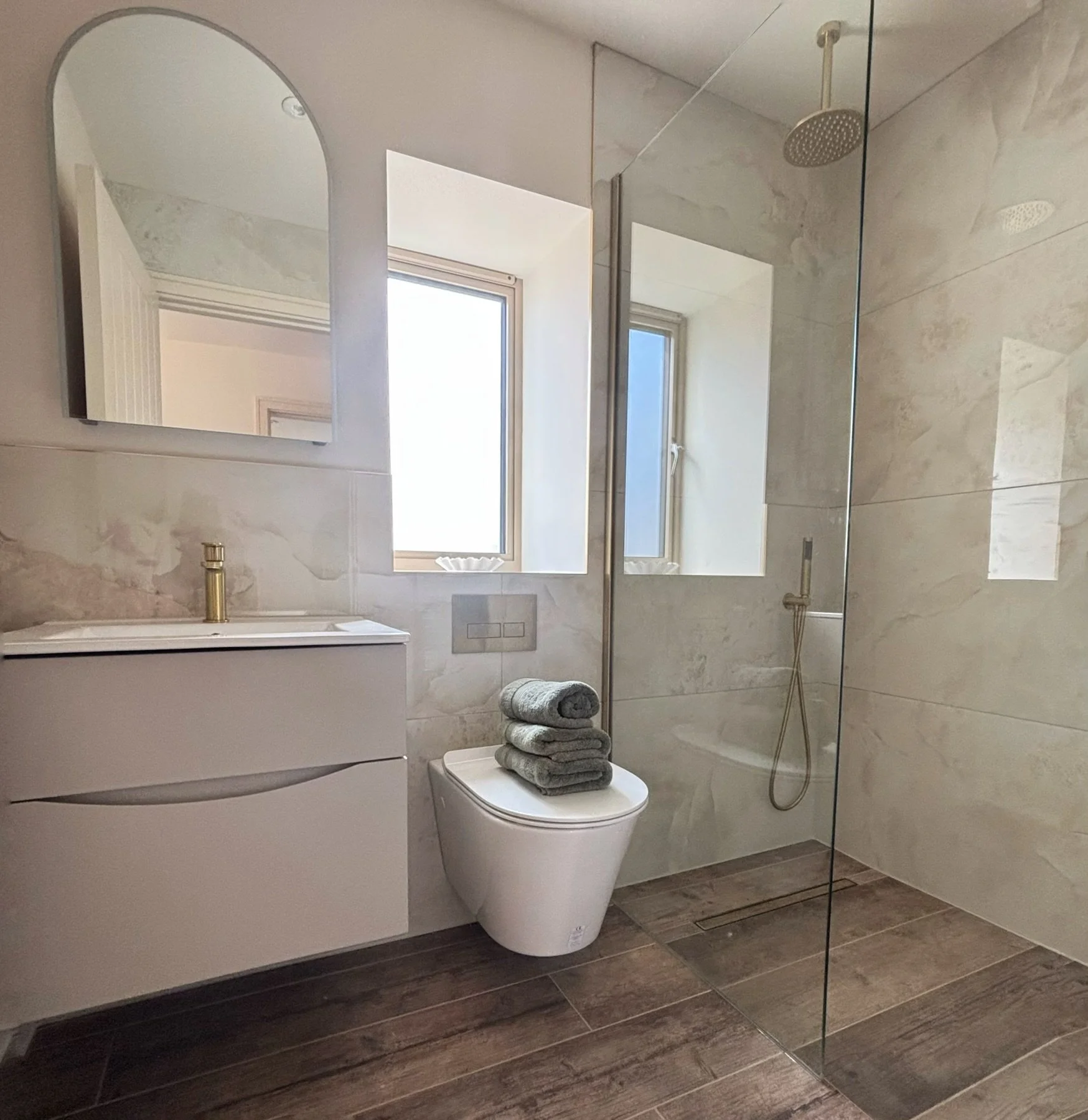 Modern bathroom with a large mirror, window, toilet with folded towels on top, shower with overhead and hand-held showerheads, marble walls, wooden floor, and a glass partition.