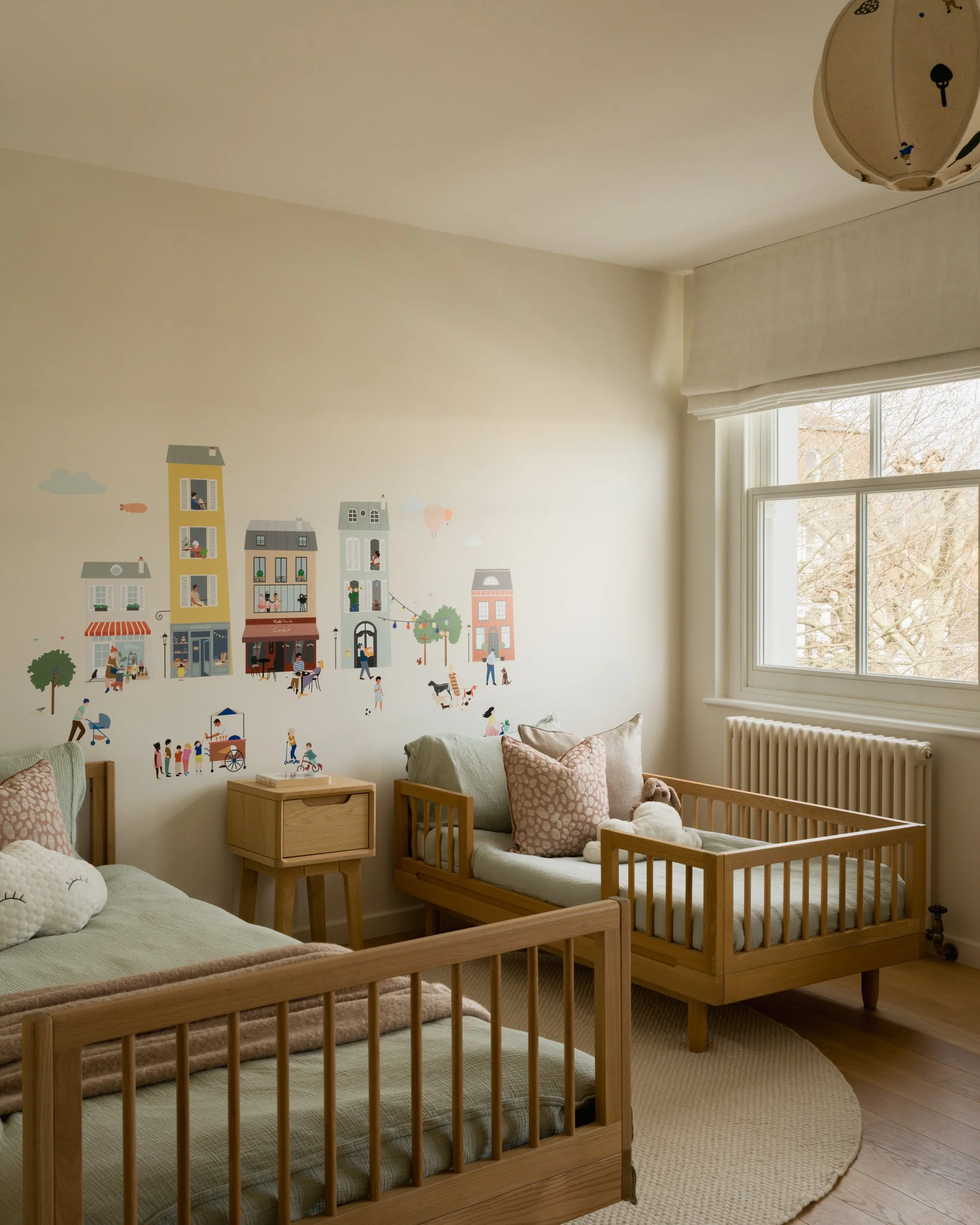 A nursery with two wooden cribs, one with a plush animal, a small wooden nightstand, a cream-colored rug, and a wall decorated with illustrated buildings, trees, and people, near a large window with curtains, and a ceiling light fixture.