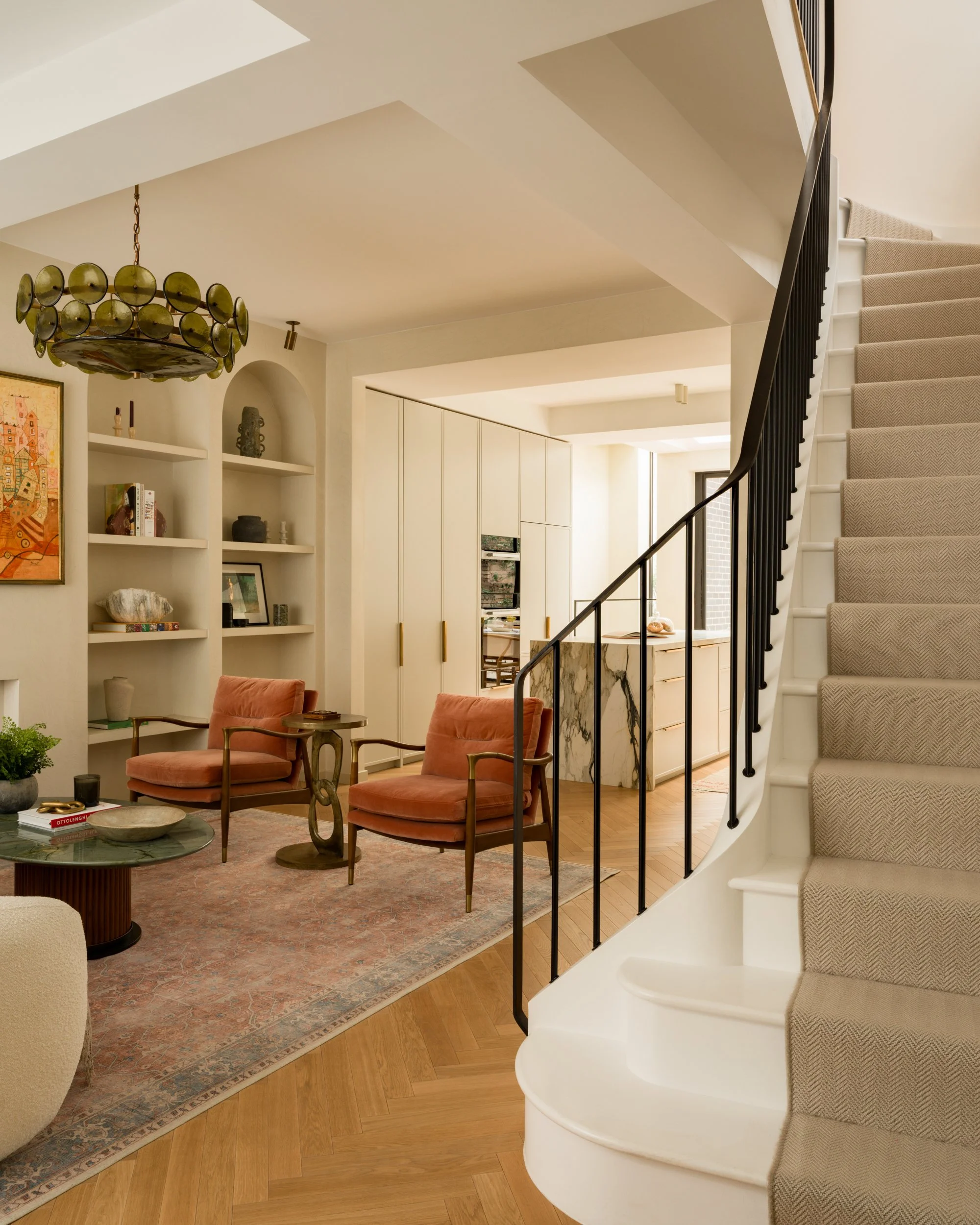 Interior view of a modern living space featuring a staircase with a beige carpet runner, a round glass coffee table, two pink armchairs, built-in white shelves with decor, and a contemporary chandelier. The background includes a kitchen area with white cabinetry and some artwork.
